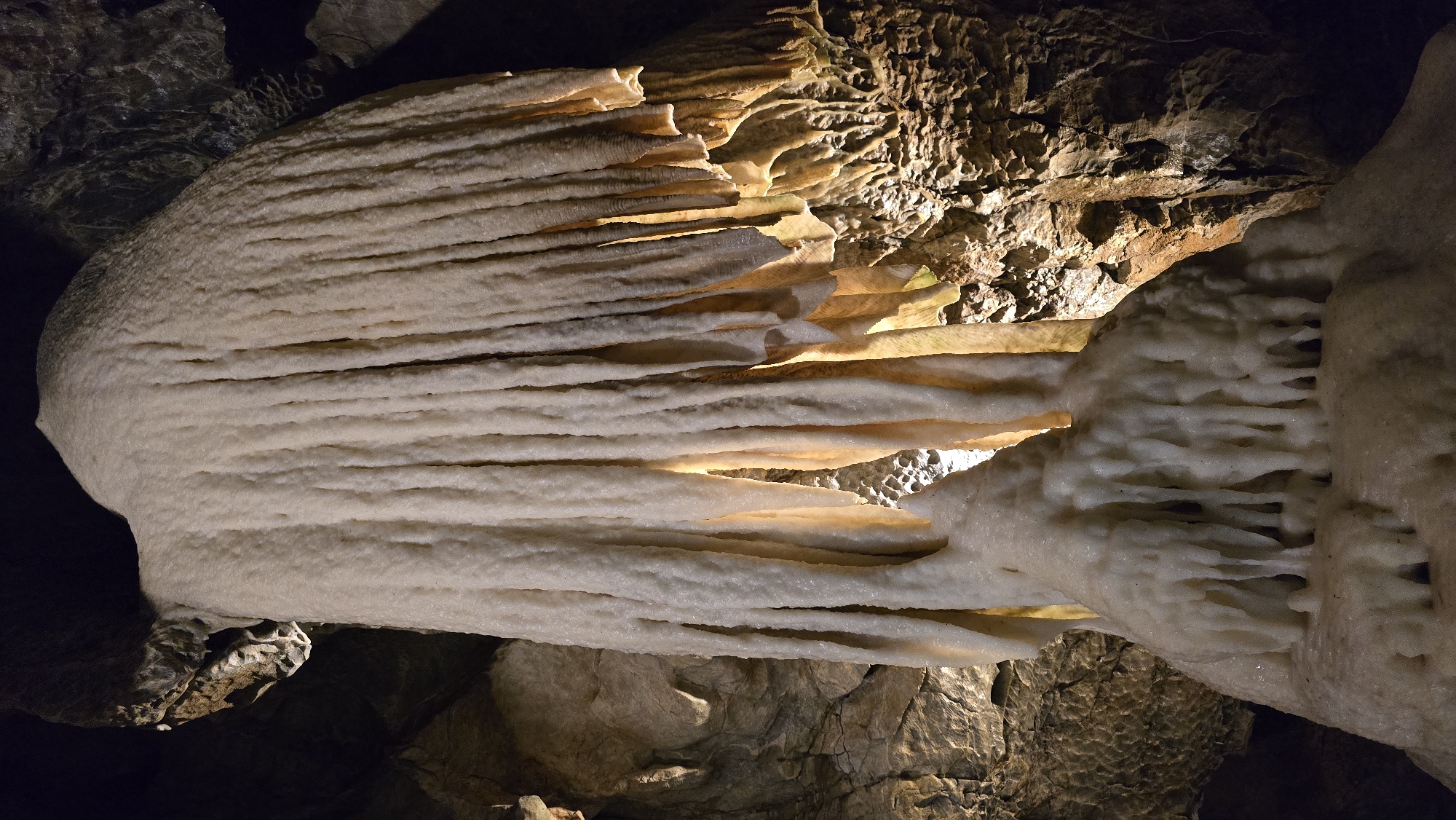 This media file is a close-up, landscape photograph of cave formations, likely stalactites and stalagmites, within a cave. The formations are predominantly white and cream-colored, with some appearing smooth and others more textured. Light, likely artificial, illuminates the formations from behind and within, highlighting their layered structures and creating a warm glow in some areas. The surrounding cave walls and ceiling are visible in the background, appearing dark, rough, and rocky, with patches of light reflecting off their surfaces. The image is shot from a low angle, looking upwards at the formations. There is no text visible in the image. The setting suggests a natural, underground environment. The texture and patterns of the mineral deposits are the primary focus, showcasing the intricate beauty of these geological formations. The lighting creates a dramatic and somewhat mysterious atmosphere. The location is identified as Vavřinec, Czechia, implying a specific geological site known for its cave systems.