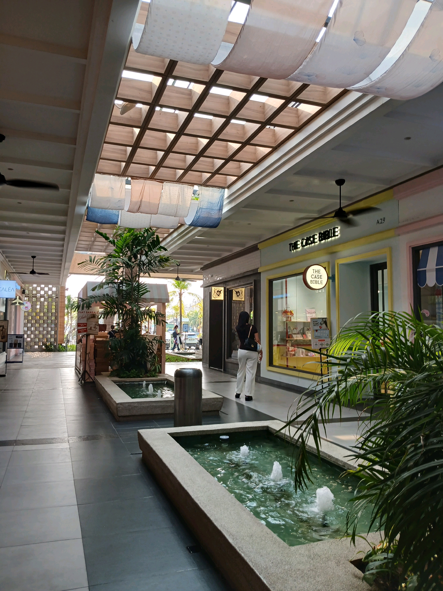 The image depicts an open-air walkway within a commercial setting, likely a shopping mall or plaza, in North Jakarta, Indonesia. The walkway features a slatted wooden roof structure with translucent panels allowing natural light to filter through, draped with decorative fabric. Along the sides of the walkway are storefronts. One prominent storefront has a sign that reads "THE CASE BIBLE." In the foreground, there are rectangular planters containing small fountains, surrounded by lush green plants. A person wearing white pants and a dark top is walking away from the camera towards the right, and other individuals are visible in the distance near the storefronts and beyond. Ceiling fans are visible in the upper portions of the walkway. The flooring is a mix of light and dark grey tiles.