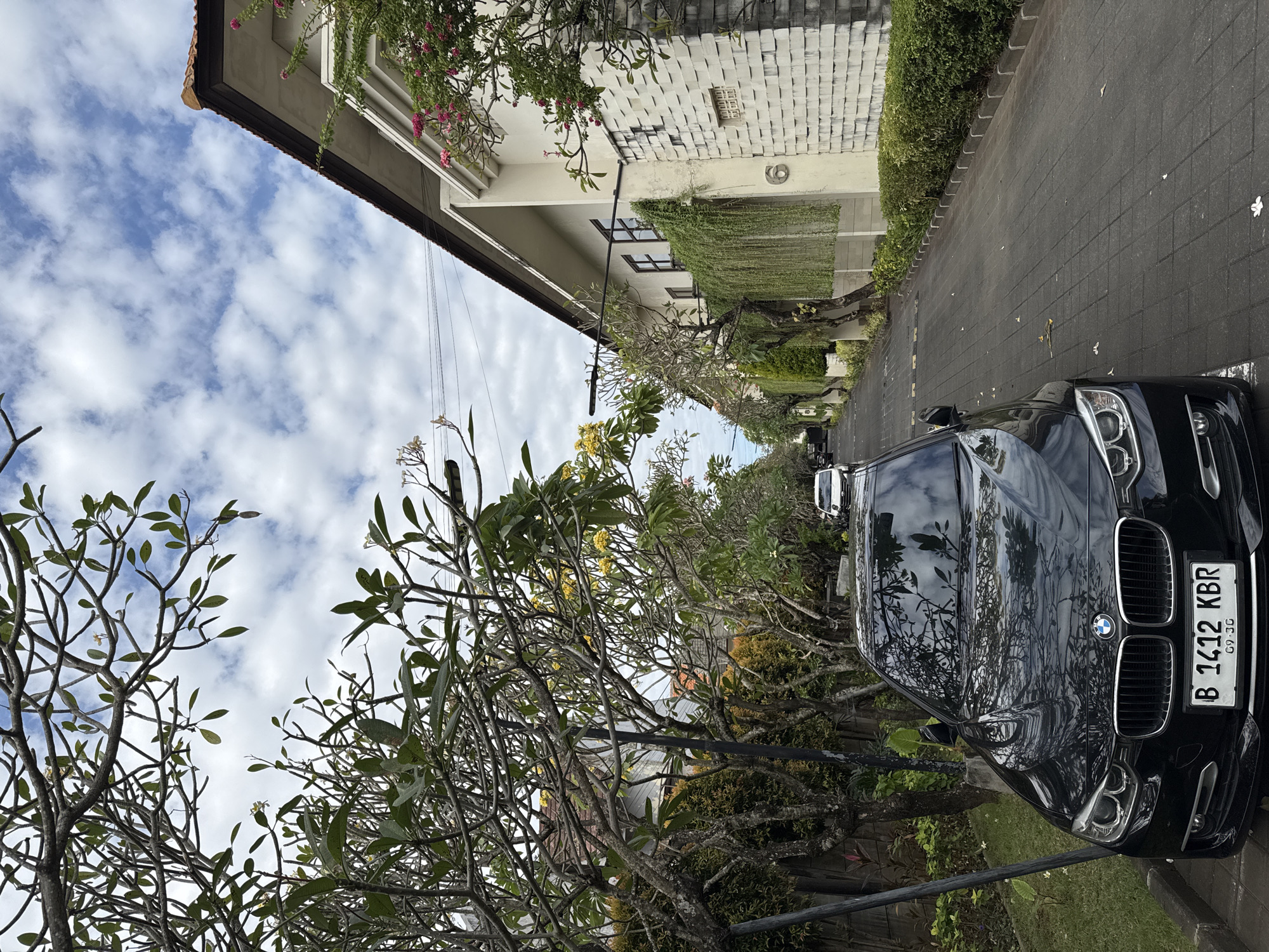 A black BMW sedan is parked on a paved driveway, its front end facing the viewer. The car has a license plate that reads "B 1412 KBR 09.26". In the background, a street runs between two buildings, one modern with white, textured walls and lush green hanging plants, and another with a tiled roof. Trees with delicate yellow flowers and green leaves line the street, casting shadows on the pavement. The sky above is blue with scattered white clouds. The scene appears to be in a residential area in Denpasar, Indonesia. There are no people visible in the image.