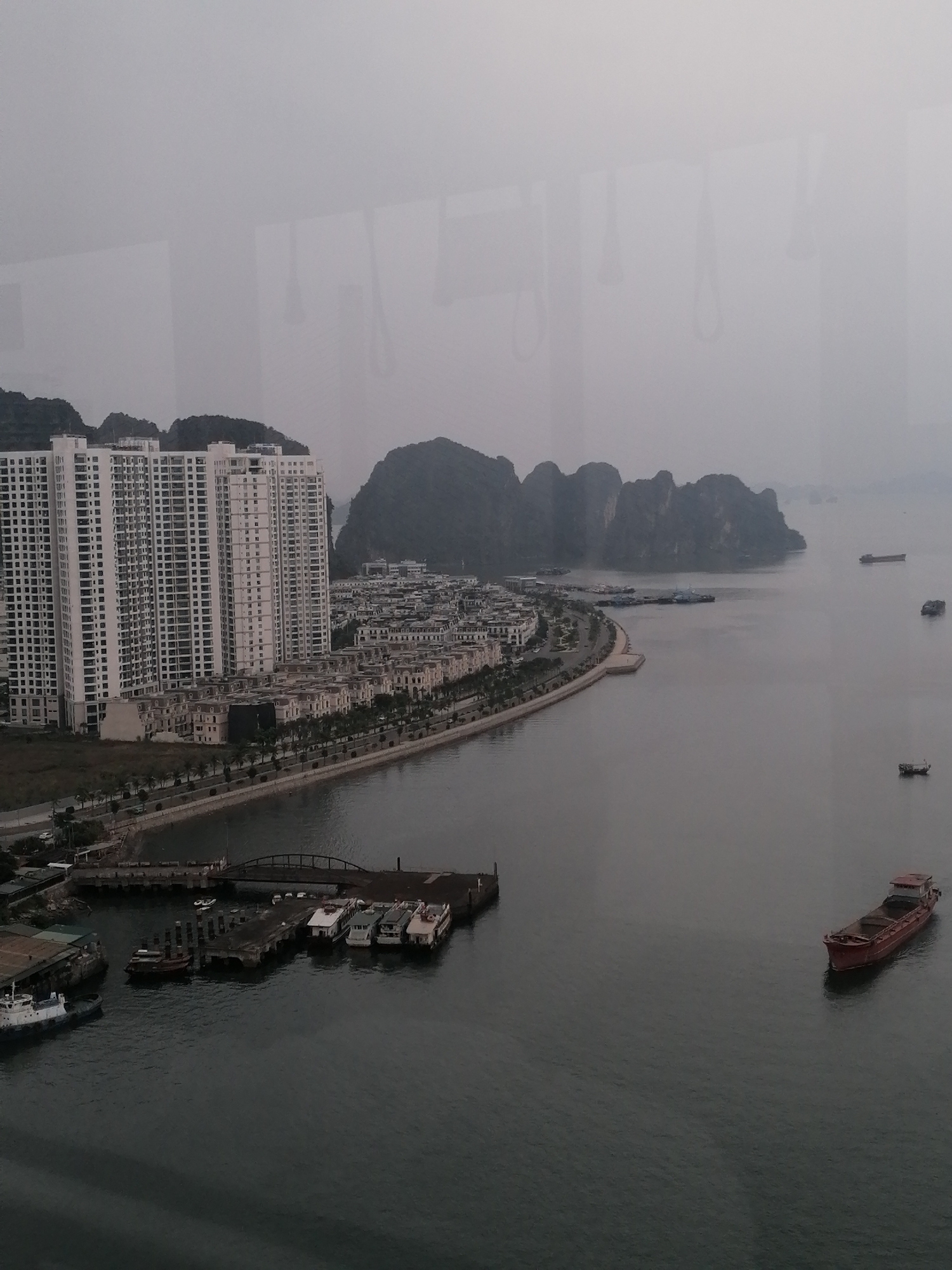 The photo captures a view of Hong Gai Ward, Vietnam, characterized by a misty or overcast atmosphere. In the foreground, a cluster of high-rise buildings dominates the left side of the frame, suggesting a densely populated urban area. Adjacent to these buildings are smaller, lower-rise structures extending along the coastline. A paved walkway lined with trees runs parallel to the shoreline, offering a pedestrian route along the water's edge. A bridge is visible, crossing a waterway near a dock. Ships and boats are docked at the pier, while others float in the open water. The background features rocky formations rising from the sea, typical of the Vietnamese coastline. The overall tone is muted due to the weather conditions, which obscure distant details. The photo is taken from an elevated perspective, possibly from a high floor of a building, overlooking the ward.