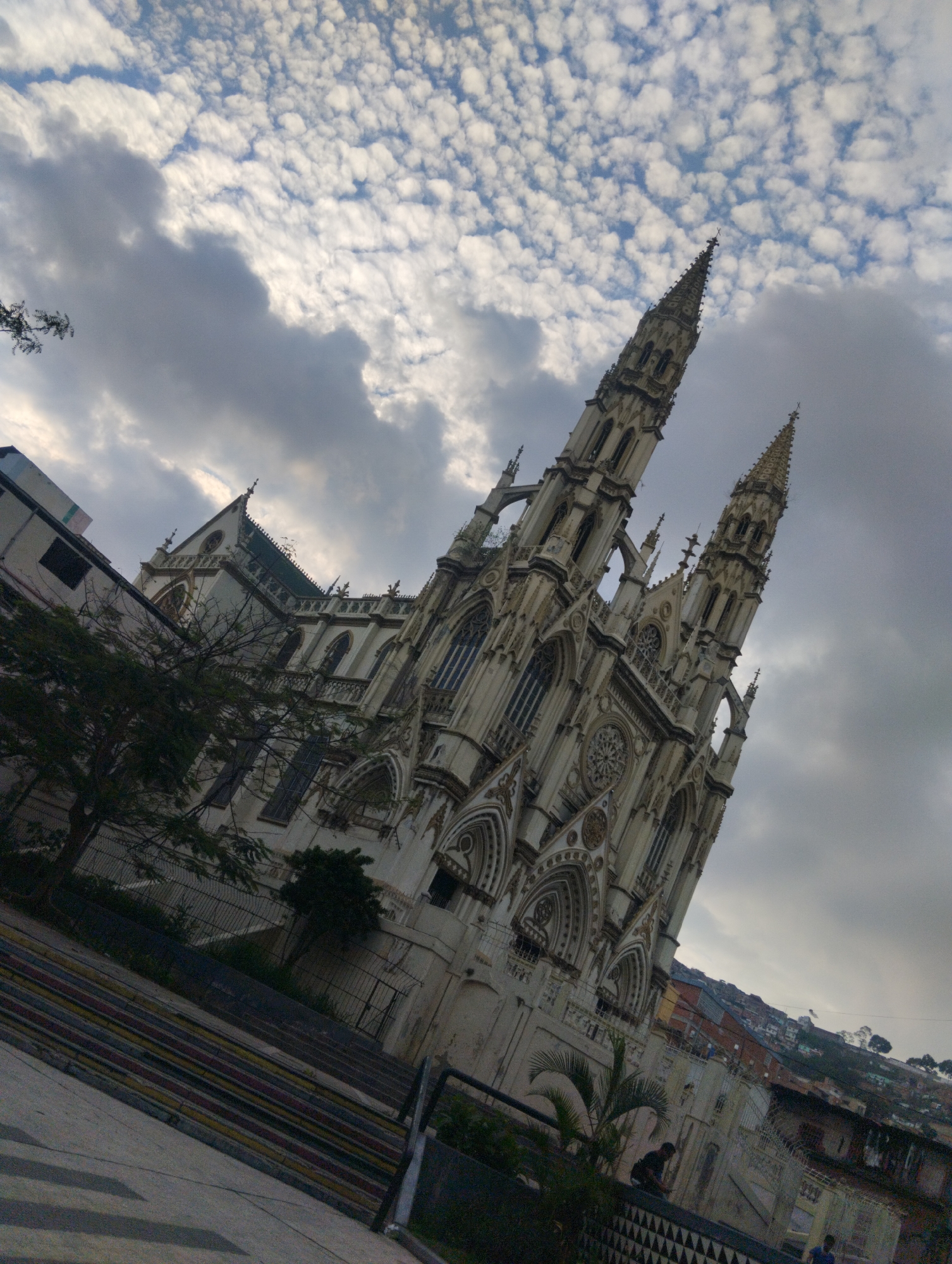 A slightly tilted medium shot depicts the Iglesia Nuestra Señora de Coromoto, a Catholic church in Unknown, Unknown. The sky is partly cloudy with a mix of white and gray clouds. The church is a tall, light-colored building with a Gothic-style architecture, featuring pointed arches and spires. In the foreground, there is a series of striped steps and a metal fence. Vegetation is visible on both sides of the frame, including trees and shrubs. Behind the church, a cityscape stretches into the distance, showing houses on a hillside. There are a few people visible near the base of the church.