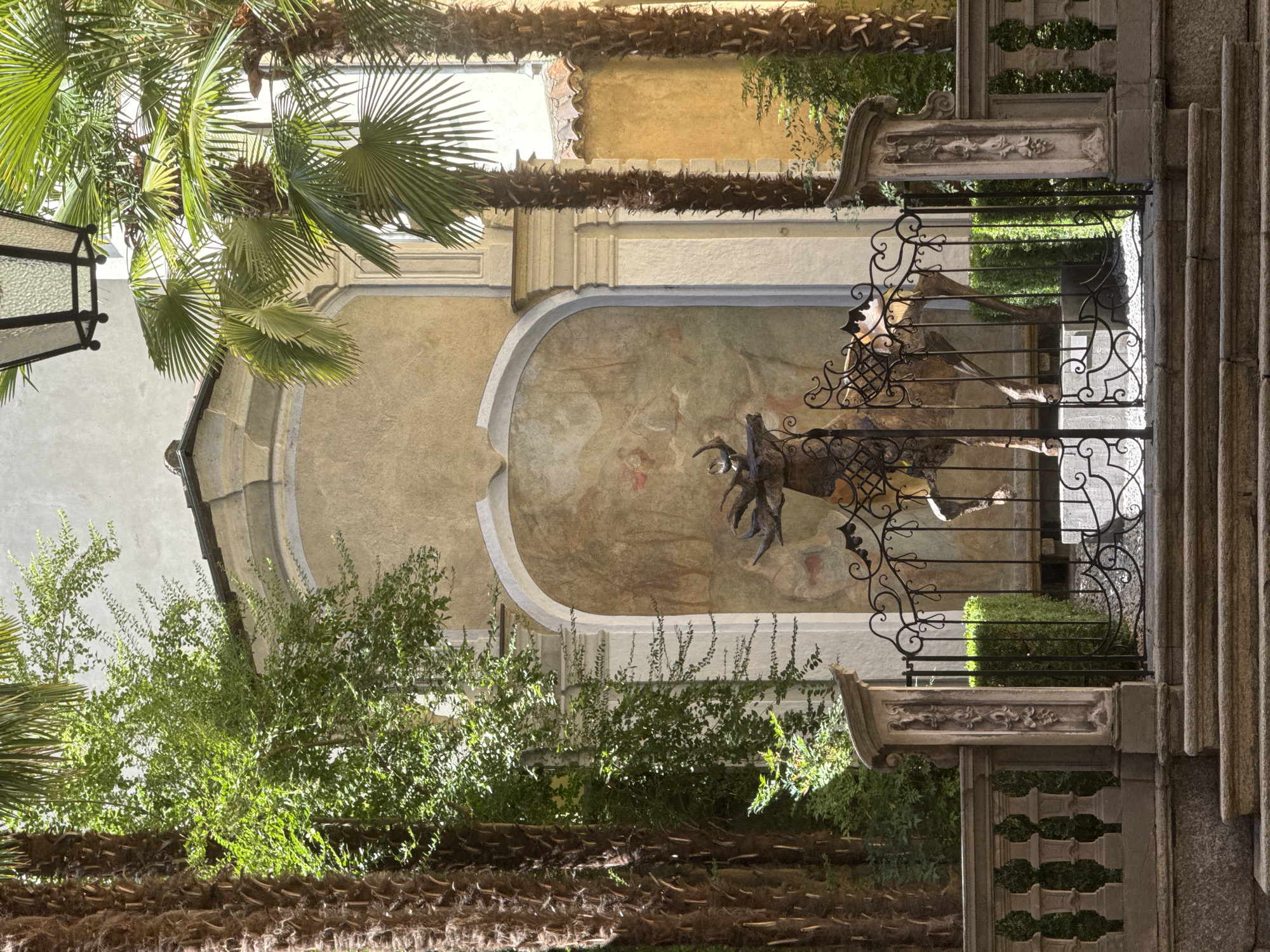 The image depicts an outdoor courtyard in Lugano, Switzerland, featuring a weathered mural behind an ornate black metal gate. The mural, visible through the gate, shows a pastoral scene with figures and animals. In the foreground of the gate, a bronze statue of a deer with large antlers stands prominently. The courtyard is flanked by lush green trees, including tall palm trees with textured trunks. Stone steps lead up to the gate, which is framed by stone balustrades and decorative pillars. A vintage lantern hangs overhead, adding to the ambiance of the scene. The lighting suggests a sunny day, with dappled sunlight filtering through the foliage.