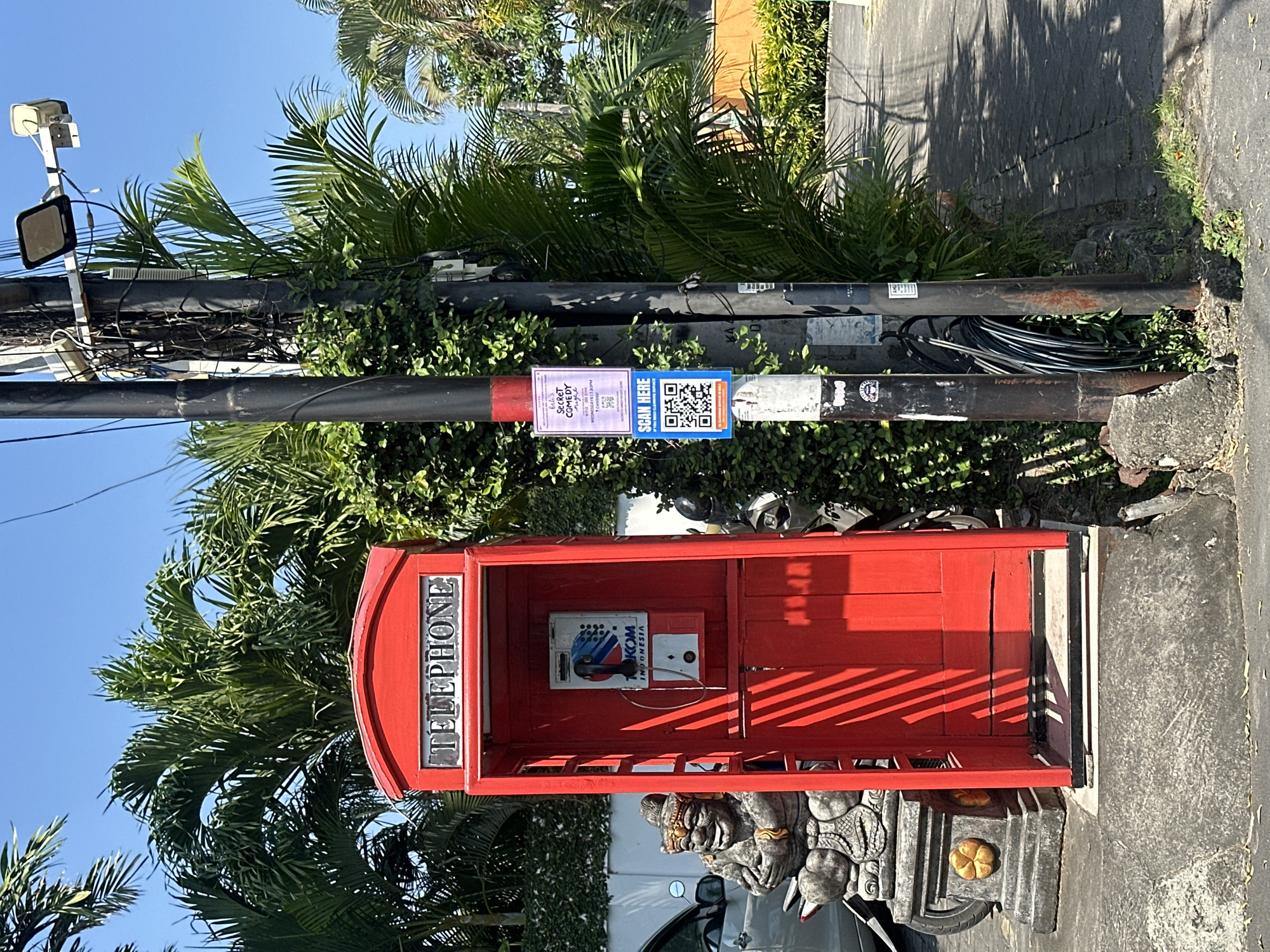 The image depicts a red British-style telephone booth situated on a paved walkway in a tropical environment, likely North Kuta, Indonesia. The telephone booth is open, revealing a retro-style telephone with a "TELKOM INDONESIA" logo. To the left of the booth, a carved stone statue of a Balinese ogre or demon sits on a plinth, partially obscuring a parked motorcycle. A car is visible in the background, also parked. Two utility poles stand to the right of the telephone booth. One pole has a light fixture and a security camera mounted at the top, and several posters and QR codes are affixed to it, including one advertising "SECRET COMEDY NIGHT" and another saying "SCAN HERE". The other pole is bundled with black cables and partially covered in green vines. The surroundings are lush with palm trees and dense green foliage, indicating a warm, sunny climate. The ground is paved with a combination of concrete and interlocking bricks. The sky is clear and blue.