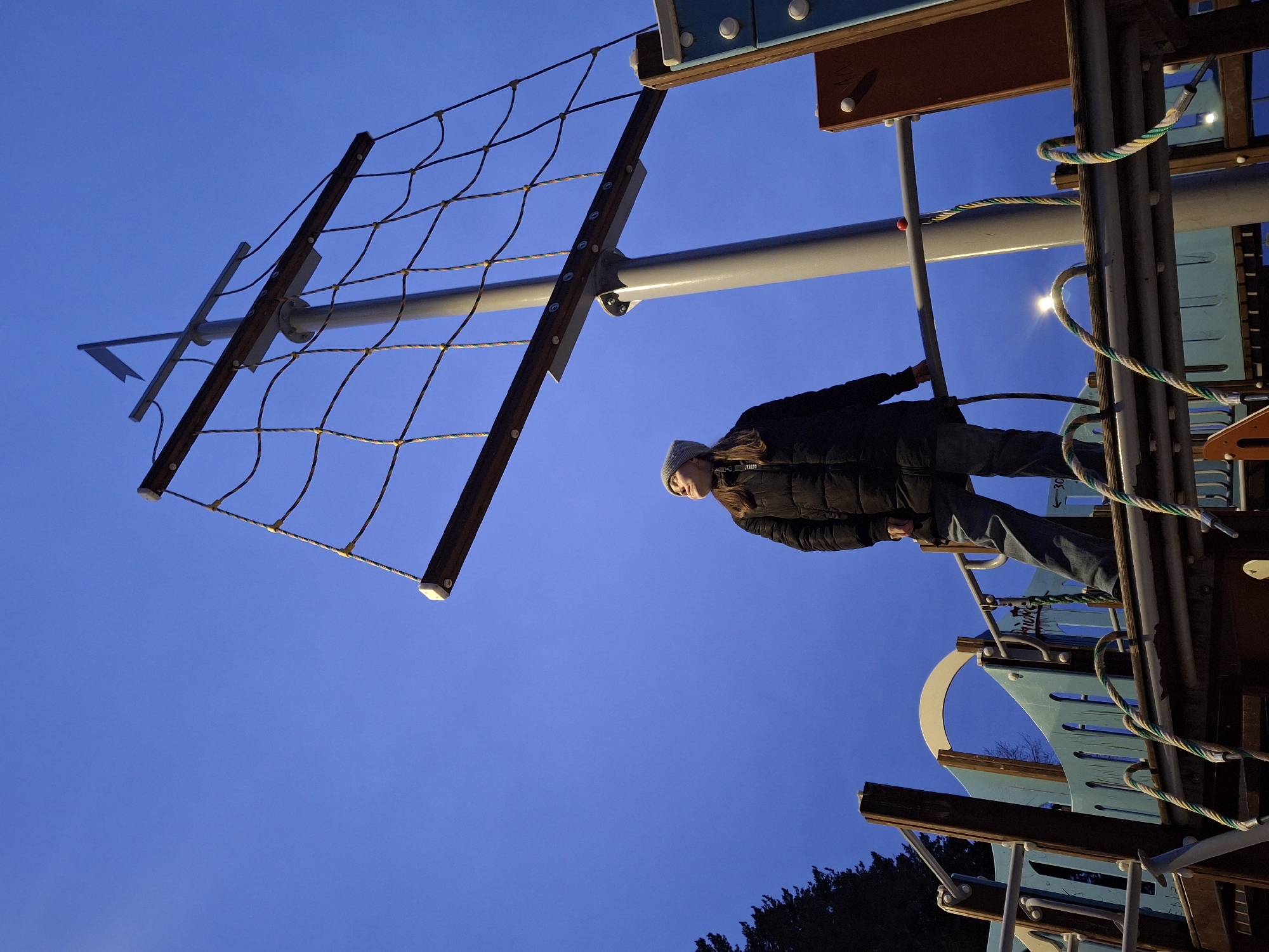 A low-angle shot depicts a woman in a black puffer jacket, jeans, and a gray beanie on playground equipment that resembles a ship's mast with a rope net, against a twilight blue sky. The playground has wooden and blue metal structures, and colorful ropes are visible. The lower right shows the top of trees. The location is Chaikovsky, Russia.