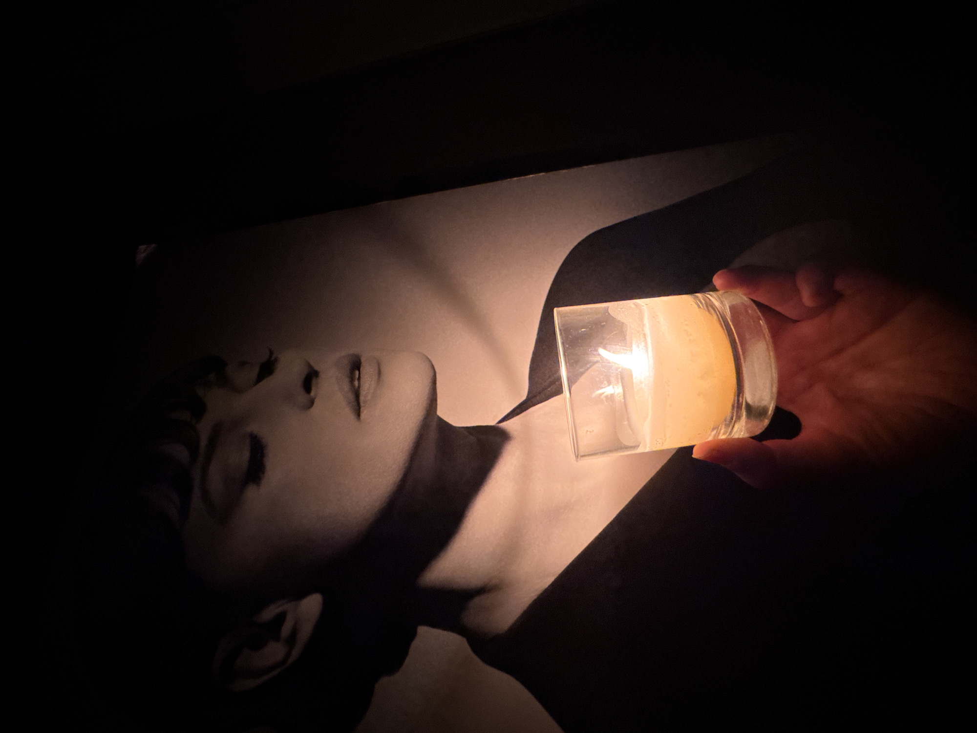 A person holds a lit candle in a glass container over a black and white image of a woman lying down. The light from the candle illuminates the woman's face and neck in the photograph. The background is dark. The location in Sofia, Bulgaria is unknown.