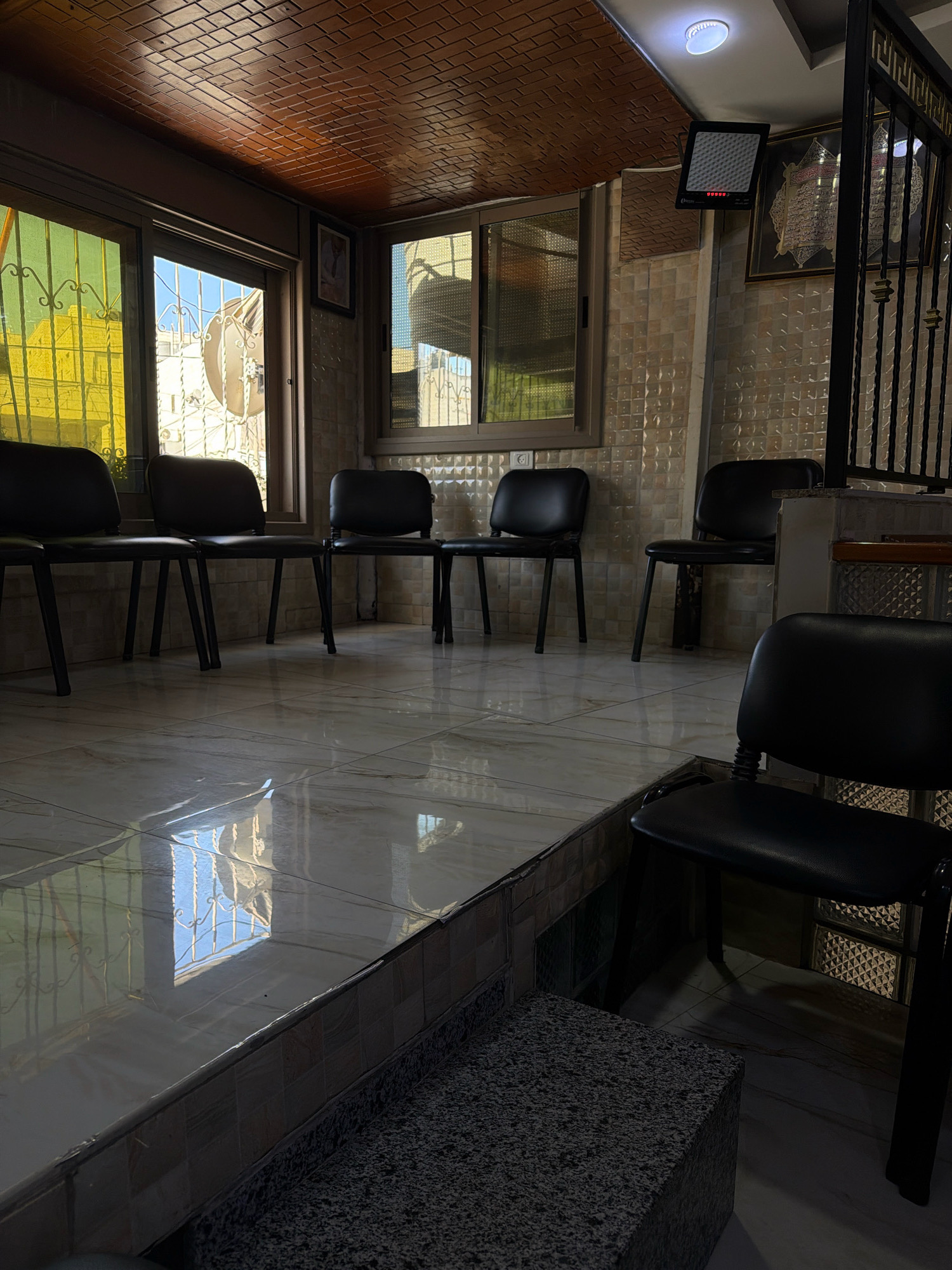 The image depicts an indoor waiting area, likely in a public building, in the city of Hebron, Palestinian Territories. The room is sparsely furnished with several black, office-style chairs arranged in rows. The floor is tiled with large, glossy white tiles that reflect the light from the windows and ceiling fixture.
The walls are covered in textured, light-colored tiles. The windows have yellow-tinted glass and metal grates on the outside, suggesting a level of security or privacy. Sunlight streams through the windows, casting reflections on the floor.
On the wall, there is a framed Islamic calligraphy artwork and a digital display device, possibly for information or announcements. A ceiling light fixture illuminates the space. A black metal gate is visible on the right side of the image. The overall impression is one of a functional, unadorned waiting space.