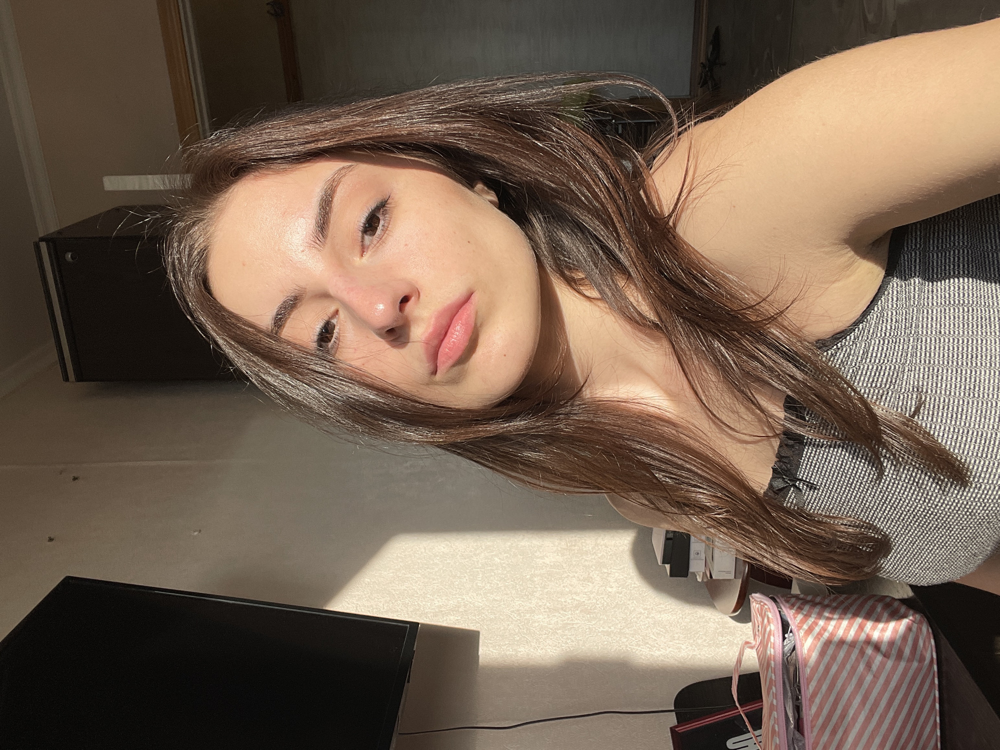 The image captures a close-up, chest-up view of a young female subject with long, dark brown hair, styled with a side part. She is wearing a grey and white patterned sleeveless top with a black lace trim at the neckline, featuring a small black bow. Her expression is neutral with a slight pout as she looks directly towards the camera. Strong directional sunlight from the left illuminates her right side and casts a distinct rectangular patch on the textured, light-colored wall behind her. The remainder of the indoor environment is primarily in shadow. To the upper left, a dark flat-screen television is mounted on the wall, with a black cable hanging vertically. Below the television, a pink and white striped open bag or container rests on a surface, beside a partially visible small, round table topped with several stacked objects. In the upper right background, a dark, tall cabinet is partially visible, with a ceiling molding above it. The scene is located in an interior room within the city of Moscow, Russia.