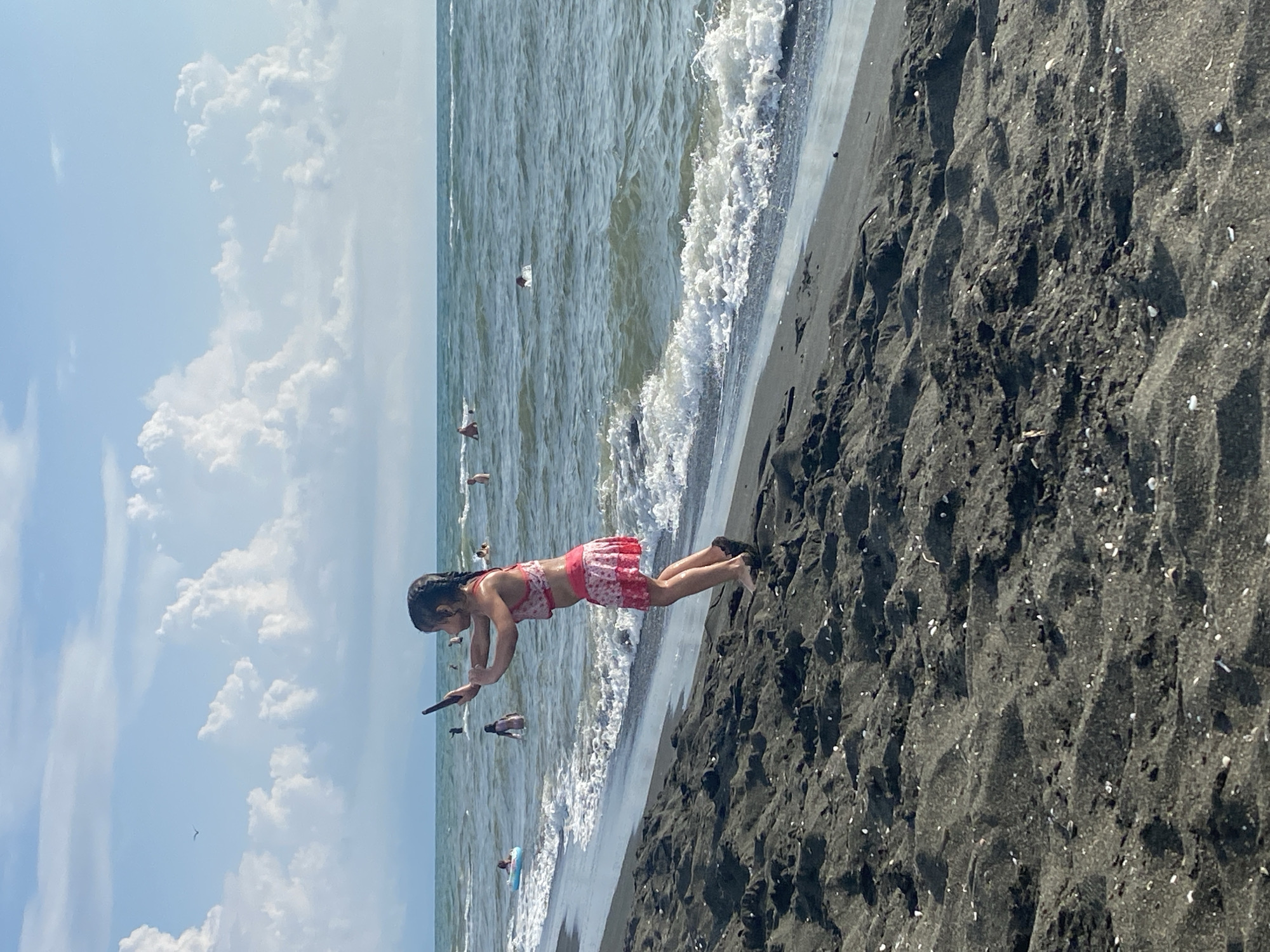 This is a vertical, eye-level, full shot of a young girl in a pink and white patterned swimsuit mid-dive into the ocean. She is on a beach of dark, coarse sand, with ripples carved into it by the tide. Her body is horizontal, angled towards the water, with her arms extended forward and her legs bent slightly behind her. She appears to be holding a dark object in her right hand, possibly a phone.
The ocean is a greenish-blue with moderate waves crashing onto the shore, creating white foam. In the background, several other people are visible in the water, some swimming and one in a blue inflatable ring. The sky is bright blue with scattered white, puffy clouds. The sun is high, suggesting it is midday. The scene is one of a casual day at the beach, likely a warm and sunny day. The girl's posture suggests excitement and playfulness. The location is Ureki, Georgia, known for its black sand beaches. There is no visible text in the image.