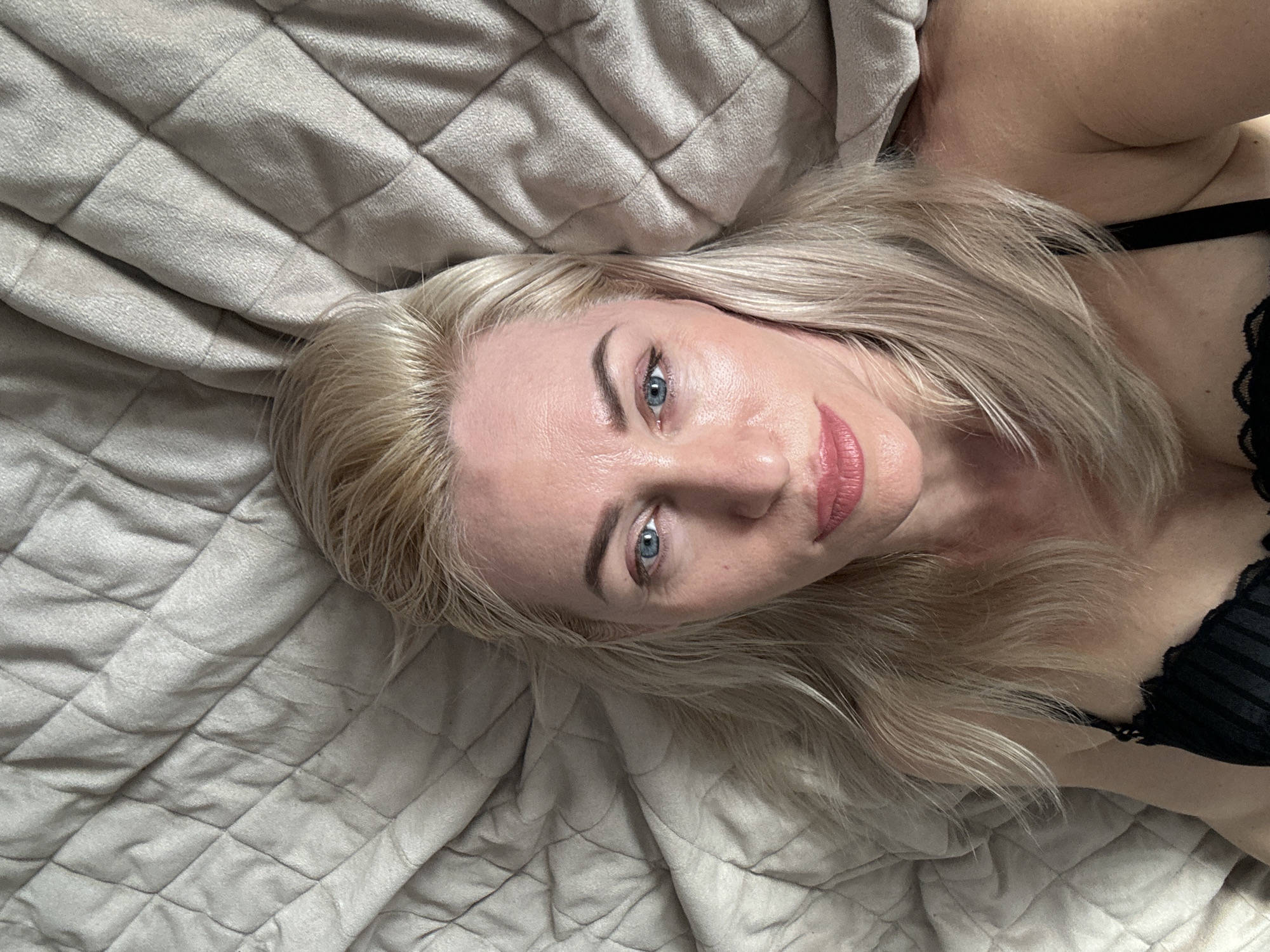 A woman with blonde hair and blue eyes is lying on a quilted grey bedspread. She is wearing a black bra with lace trim. The location appears to be indoors, but the city and specific location are unknown.