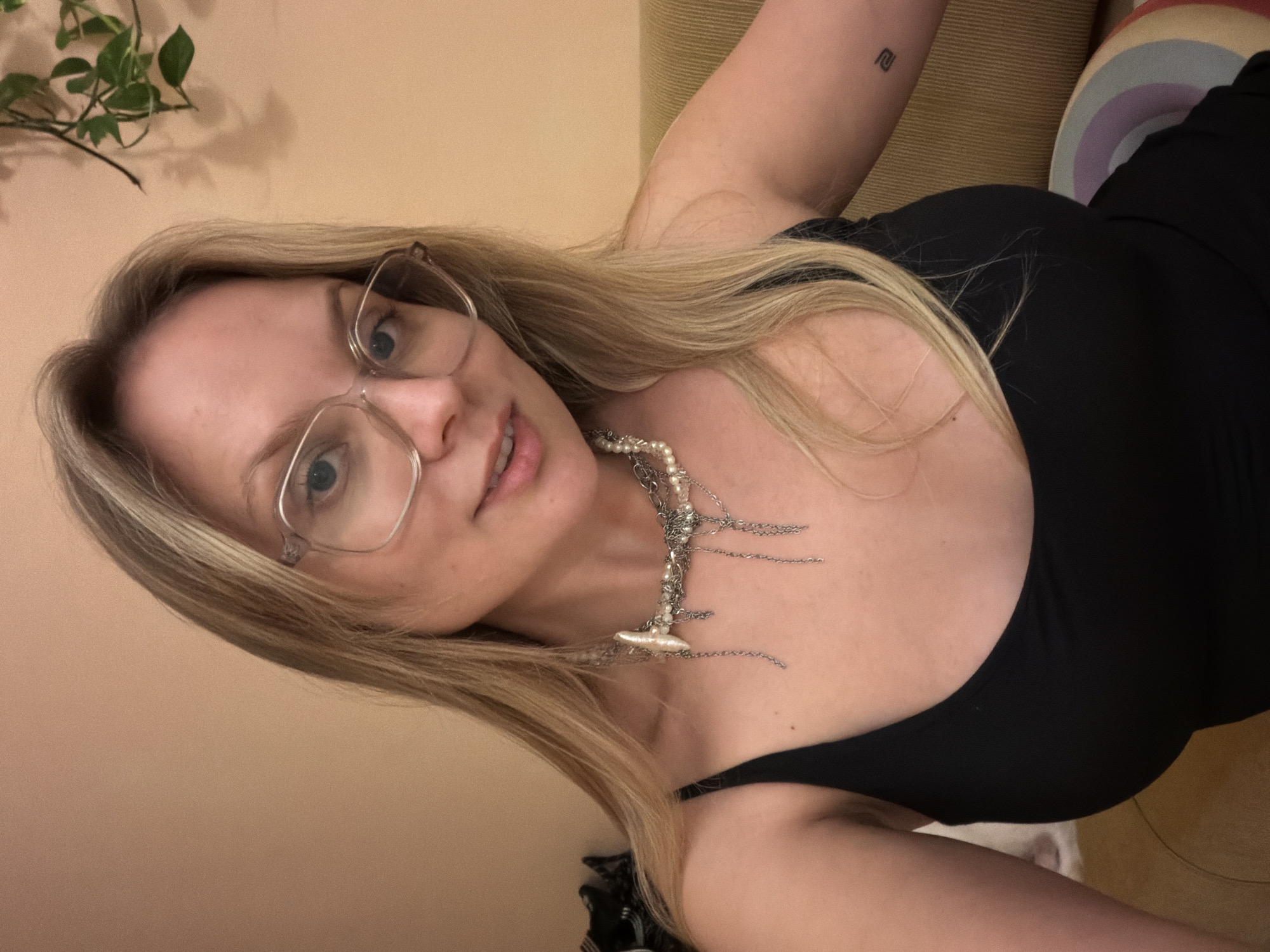 A blonde female subject with light-colored eyes and clear-framed eyeglasses is depicted in a close-up, head-and-shoulders portrait. She is wearing a black scoop-neck tank top and a multi-strand necklace composed of irregular pearls, silver-toned chains, and a prominent elongated baroque pearl or shell element. A small, dark, abstract tattoo resembling a stylized number '2' or 'S' is visible on her right forearm/bicep. Her lips are slightly parted in a subtle smile, and she maintains direct eye contact with the camera. The background features a plain, light peach-colored wall. In the upper right, a green vine plant with several leaves is visible, casting a faint shadow. Sections of textured soft furnishings, including light green and tan cushions, are present on the right and lower left sides of the frame. A round, decorative cushion with rainbow stripes (red, orange, yellow, green, blue, purple) is visible in the lower right. The scene is illuminated by warm, indirect light, characteristic of an indoor setting. This scene is understood to be located within the city of Tel-Aviv, Israel.
