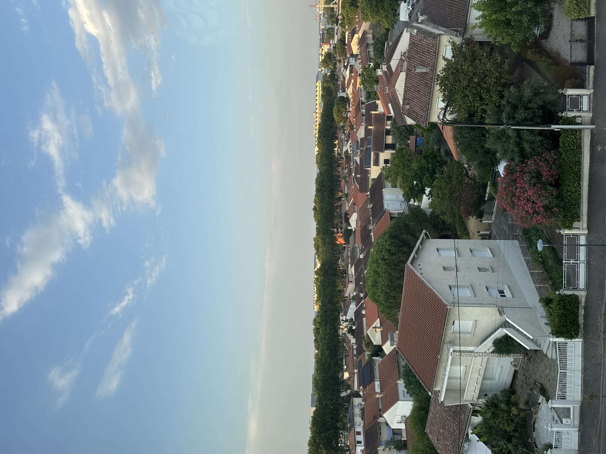 The image depicts a suburban residential area in Colomiers, France, on a clear day with scattered clouds. The scene is an aerial view looking down on a collection of houses with red-tiled roofs. The architecture suggests a typical European suburban neighborhood.
In the foreground, a light-colored house with a red roof is prominently featured. It has multiple windows and a balcony on the upper level. A staircase leads up to the entrance. The house is surrounded by greenery, including trees and bushes. A white metal fence and gate are visible in front of the property, along with a paved driveway.
Further back, more houses with red roofs are densely packed, interspersed with trees. A row of mature trees forms a green line across the middle ground, separating the immediate neighborhood from structures in the distance.
In the background, larger buildings, possibly commercial or industrial, are visible behind the tree line. A red construction crane is also present in the far distance, suggesting ongoing development or infrastructure projects. The sky is blue with streaks of white clouds, indicating fair weather. The overall impression is one of a peaceful, well-established residential district within a larger urban or peri-urban landscape. No people are visible in the image.