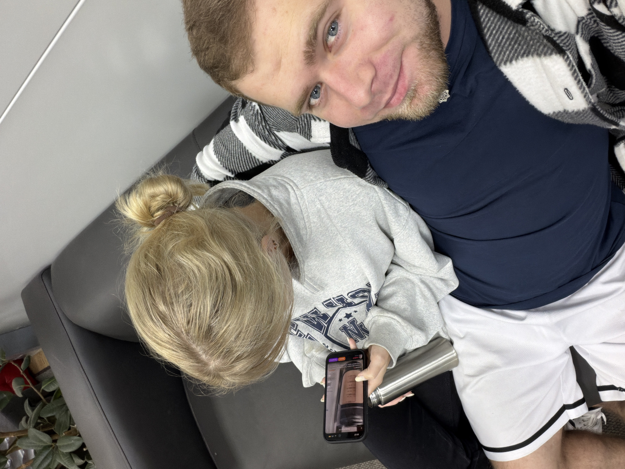 A man and a woman are reclining on a gray sofa. The man has light brown hair, a beard, and blue eyes. He is wearing a navy blue t-shirt and white athletic shorts with black stripes. A black and white plaid shirt is draped over him. The woman has blonde hair pulled back in a ponytail. She is wearing a gray hooded sweatshirt with a navy blue logo, and black pants. She is holding a black smartphone in her right hand and a stainless steel water bottle in her left hand. The image appears to be taken indoors with a white wall in the background. There is no information available in the image to confirm if it was taken in Chaikovsky, Russia.