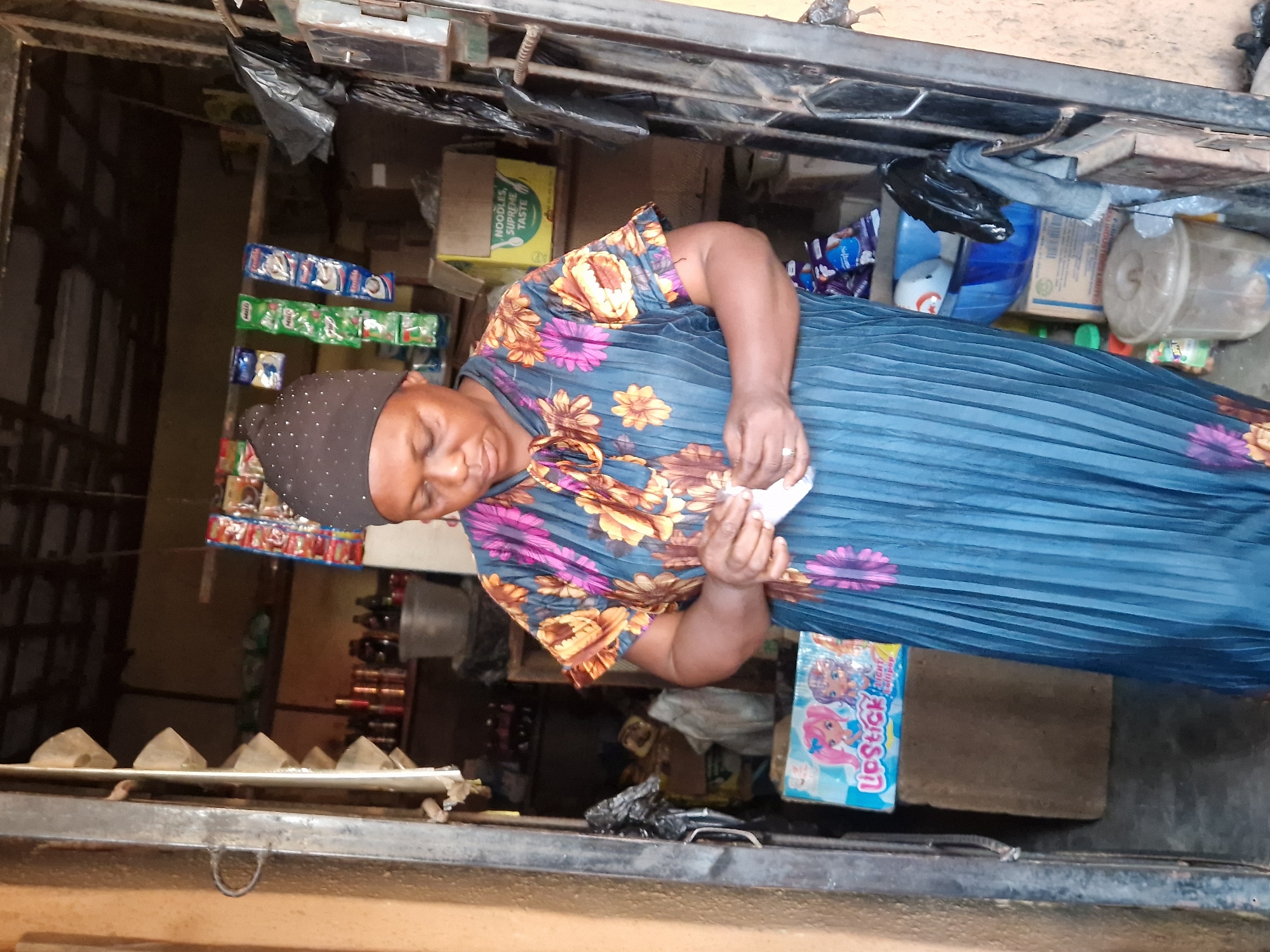 A woman wearing a dark headscarf and a floral-patterned, pleated blue dress stands in the doorway of a small shop. She is looking down and holding a small white object in her hands. Behind her, the shop is filled with various items, including packaged goods, bottles, and containers, displayed on shelves and on the floor. The shop appears to be an open-air market stall in Jos, Nigeria, characterized by its rustic construction and assortment of merchandise.