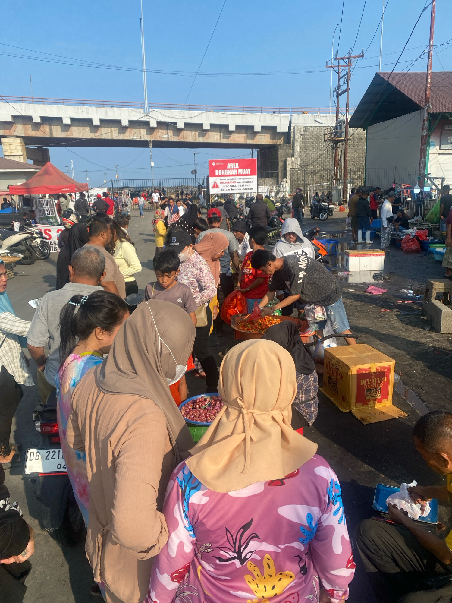 The image depicts a busy outdoor market scene, likely in Manado, Indonesia, under a bright, clear sky. A large overpass, possibly part of a highway or elevated road, dominates the background, with concrete pylons and textured concrete walls visible. Power lines and poles are scattered throughout the upper portion of the frame, suggesting an urban or semi-urban environment.
In the foreground and midground, a dense crowd of people is engaged in commercial activity. Many individuals are dressed in casual attire, with some women wearing hijabs. Several motorcycles are parked among the stalls, indicating modes of transportation. Red and white tents are set up, likely for vendors.
Merchandise is displayed on the ground and on tables. In the mid-right, a large cardboard box labeled "GUDANG GARAM" is prominent. A variety of goods, including what appears to be fruits or vegetables in large bowls and baskets, are visible. One person is interacting with a pile of red onions on a blue tray.
The ground is paved, and there are signs of recent activity, with some wet patches and scattered debris. A red banner with Indonesian text is prominently displayed on a pole in the center of the image, reading "AREA BONGKAR MUAT" and further instructions, suggesting a loading and unloading zone within the market.
The overall impression is one of lively commerce and community interaction within a functional, albeit somewhat unrefined, urban market setting.