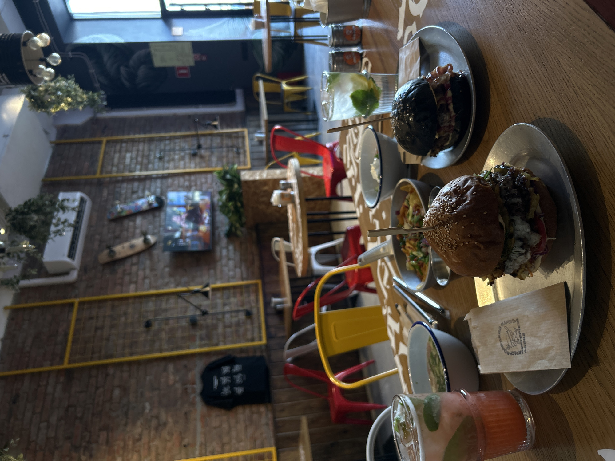 The image depicts a casual dining scene in a restaurant. The foreground is dominated by a wooden table laden with food and drinks. Two burgers are prominently displayed on metal plates. The burger on the left has a sesame seed bun and appears to be a traditional burger, while the burger on the right features a black bun and visible bacon. To the left of the burgers, there are two drinks, one in a tall glass with ice and mint, and another in a shorter glass with a pink hue and mint. There are also bowls of food, possibly sides or dipping sauces, and a small brown paper bag with text on it. The background reveals the restaurant's interior, characterized by exposed brick walls, yellow accents on scaffolding-like structures, and hanging plants. Several tables and chairs are visible, with a mix of red and yellow seating. A television screen is mounted on the wall, displaying an image. The lighting appears to be a mix of natural light from a window on the right and interior lighting. The overall atmosphere suggests a modern, trendy eatery. There are no people visible in the image.