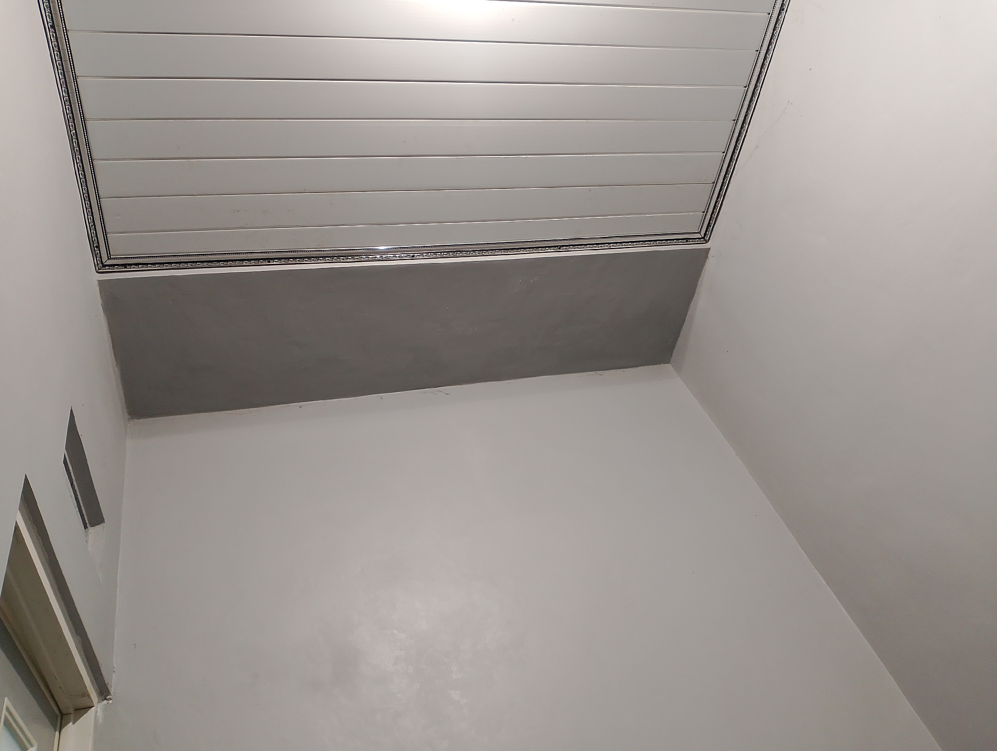 The image shows an interior view of a ceiling and upper walls of a room. The ceiling is made of white horizontal planks with a decorative border. Below the ceiling, there is a rectangular section of a plain gray surface. The walls are painted white and extend downwards from the ceiling. On the left side of the image, there is a portion of a window frame and what appears to be a ventilation grate or opening in the wall. The lighting suggests an indoor setting with artificial light sources, possibly from above or within the ceiling. This scene is a generic interior shot and does not provide specific visual cues to determine its location within Magelang, Indonesia.