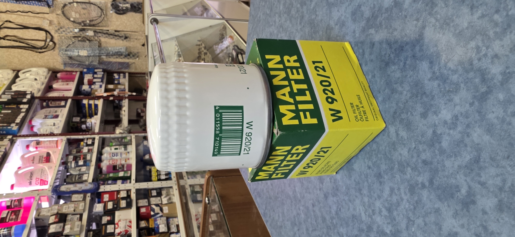 The image shows a close-up of a Mann Filter W 920/21 oil filter and its packaging, placed on a table or counter. The filter is white with a barcode and model number printed on it. The box is predominantly green and yellow, with "Mann Filter" and "W 920/21" prominently displayed. The background reveals shelves stocked with various automotive products, suggesting a retail environment like an auto parts store. The lighting is bright, illuminating the product and the shelves behind it. The scene appears to be indoors, likely within a commercial establishment in Kryvyi Rih, Ukraine.