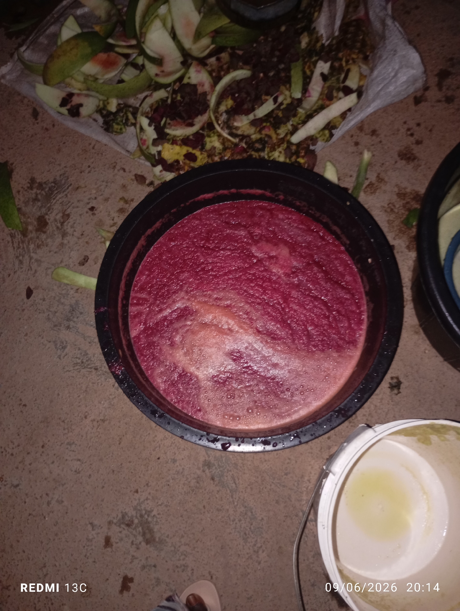 A high-angle shot shows a large black bucket filled with a thick, dark red liquid. The liquid has a frothy texture on top, indicating it may be a juice or smoothie. To the left of the bucket is a pile of discarded fruit peels, a white cloth bag, and some green stems, suggesting the processing of fruit. In the lower right corner, a white bucket with a small amount of light yellow liquid is visible. The ground appears to be concrete or dirt. The image is timestamped with "09/06/2026 20:14" and "REDMI 13C". The scene is outdoors, likely in a yard or preparation area, given the discarded materials and the rough ground. No people are directly visible, though a foot wearing a sandal can be partially seen in the bottom center of the image.