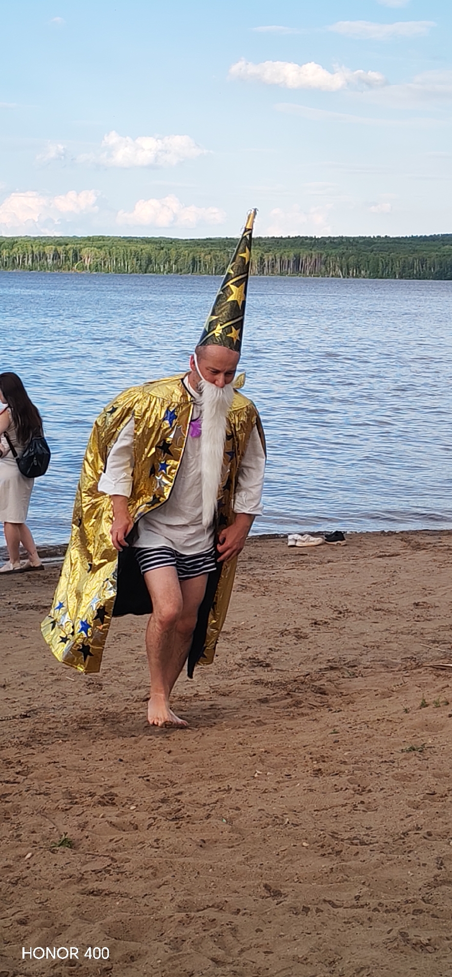 A man dressed as a wizard walks on a sandy beach next to a large body of water. He is wearing a gold cape adorned with stars, a white shirt, and striped shorts. He also has a long white beard and a tall, pointed hat with stars. In the background, there is a forest and a blue sky with clouds. On the beach to the left of the wizard, a woman in a light-colored dress is visible. Further down the beach, near the water, a pair of shoes are placed. The image also shows the watermark "HONOR 400" in the bottom left corner. This scene is likely taking place at the Kama River or a reservoir in Chaikovsky, Russia, given the presence of a large body of water and a forest.