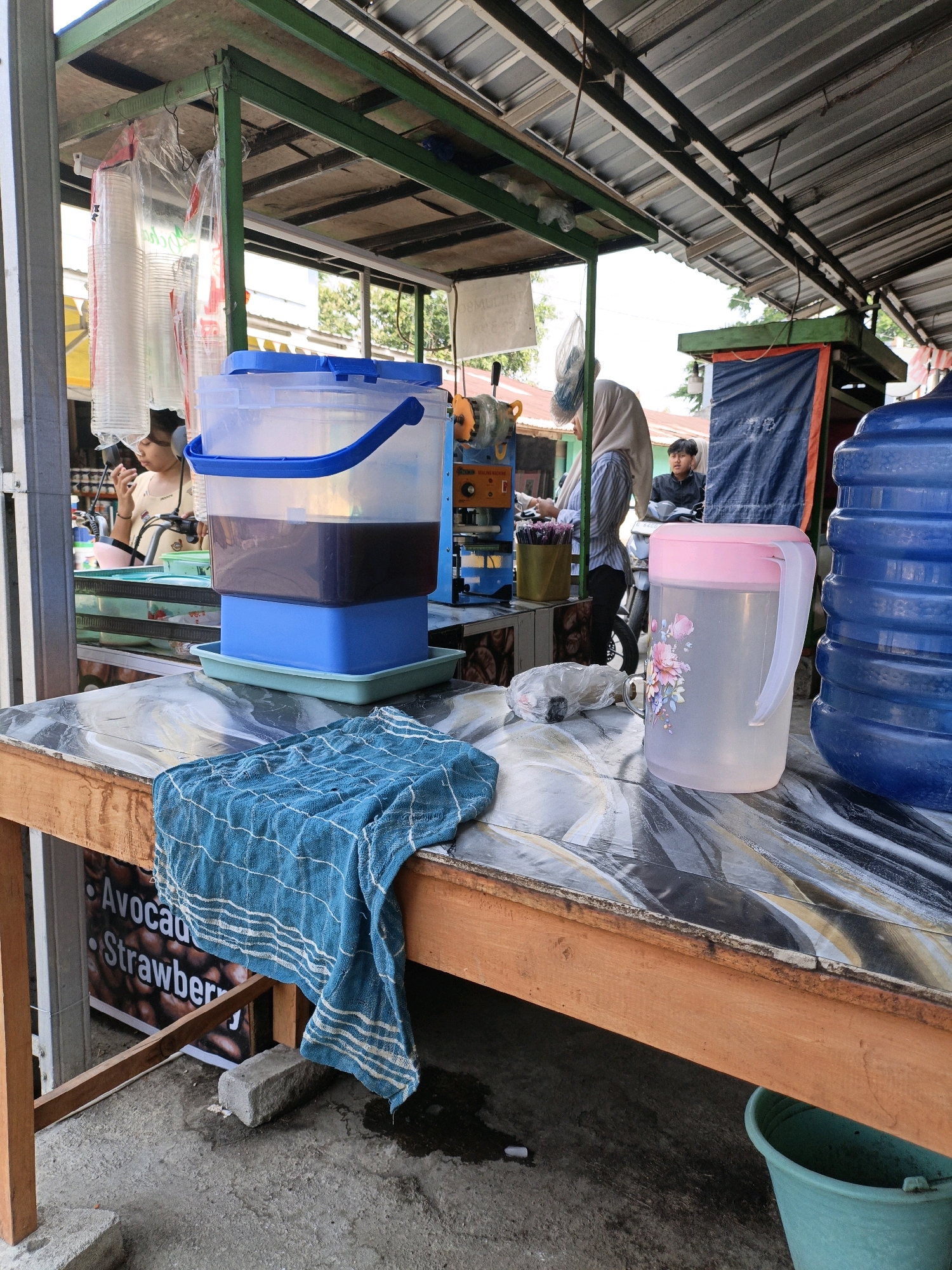 The image depicts an outdoor food and beverage stall in Tanahbaya, Indonesia, likely operating on a street or walkway. In the foreground, a wooden table is set with a blue and white checkered cloth draped over its edge. To the right of the cloth, a clear plastic pitcher with a pink lid, decorated with floral motifs, stands on the table. Further to the right, a large blue plastic water cooler jug is partially visible.
Behind the table, a large transparent plastic dispenser filled with a dark liquid, possibly iced tea or coffee, sits atop a blue plastic base and a green tray. To the left of the dispenser, a woman wearing a light-colored headscarf and a yellow shirt is partially visible, appearing to be interacting with a customer or preparing drinks. Behind her, stacks of clear plastic cups are hanging, along with some utensils. Further back, another person, a young male wearing a black jacket, is standing near a motorcycle. In the background, there's an orange and yellow beverage machine and a blue tarp hanging. The stall appears to be situated under a corrugated metal roof supported by green metal framing. The ground surface is concrete, with some puddles and a turquoise plastic bucket visible near the table's base. A sign with "Avocado" and "Strawberry" printed on it is visible on a post to the left. The scene suggests a busy commercial operation typical of Indonesian street vendors.