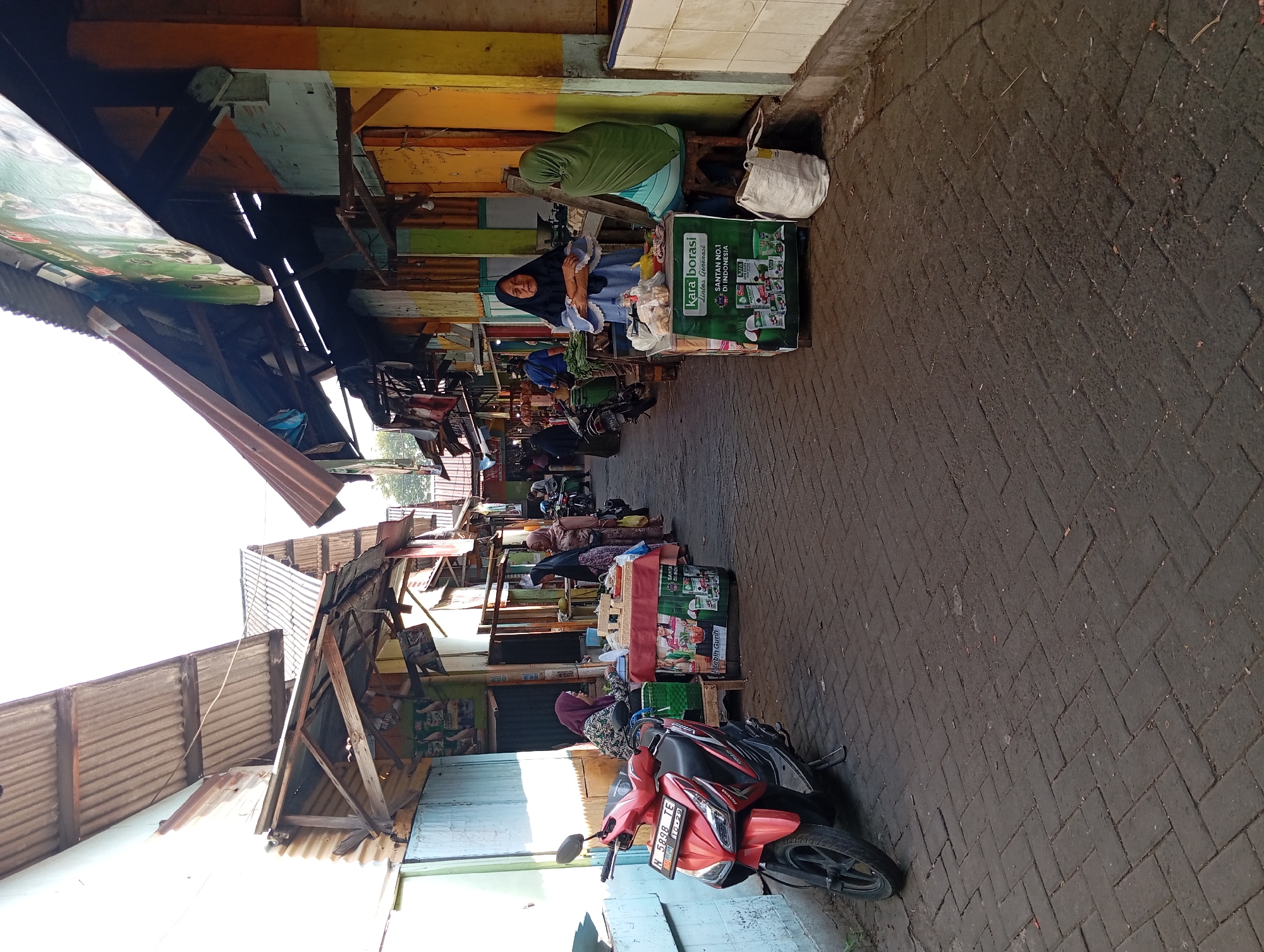 The image shows a street scene in Kepulungan, Indonesia, likely a market area. The pavement is made of dark, interlocking bricks. On the left, a red and black scooter is parked next to a light blue wall. Further down the street, several stalls are set up, some with corrugated metal roofs supported by wooden structures.
People in traditional Islamic attire, including hijabs and headscarves, are visible at the stalls. One woman wearing a blue hijab and white trim is standing behind a green display cart advertising "Kara Borasi Santan No. 1 Di Indonesia" (Kara Borasi Coconut Milk No. 1 in Indonesia). Another woman in a green hijab is seated on a stool next to this stall. Further down, another woman in a patterned hijab is seated at a stall displaying goods on a red and white tablecloth.
The buildings lining the street appear to be simple structures with colorful facades, painted in shades of orange, yellow, green, and blue. Some have signs and tarps overhead. The overall impression is of a bustling, open-air market environment.