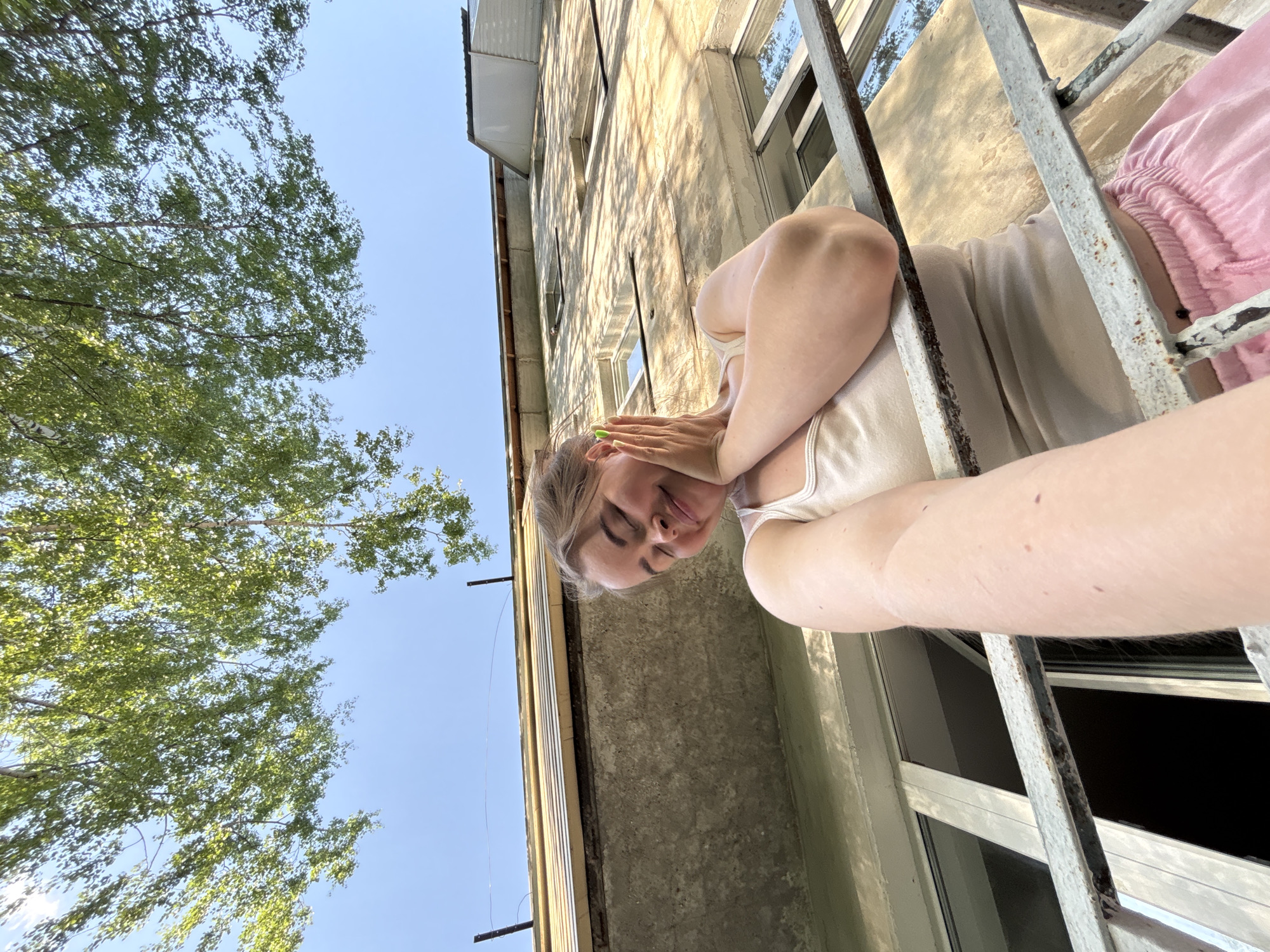 A medium shot, taken from a low angle, captures a woman posed in front of a window with a metal security grate, against a backdrop of a building and a tree. The woman is fair-skinned, with light brown hair and visible arm freckles. She is wearing a white tank top and pink shorts. Her face is tilted down, and her hands are near her face, with neon green nail polish visible.
The building behind her has a concrete facade with multiple windows, casting shadows on its surface. To the left of the frame, a tree with dense green foliage is visible against a clear blue sky.
While the location cannot be definitively determined from the image alone, based on the provided context (city of Izhevsk, Russia) the style of architecture visible is consistent with residential buildings in that region.