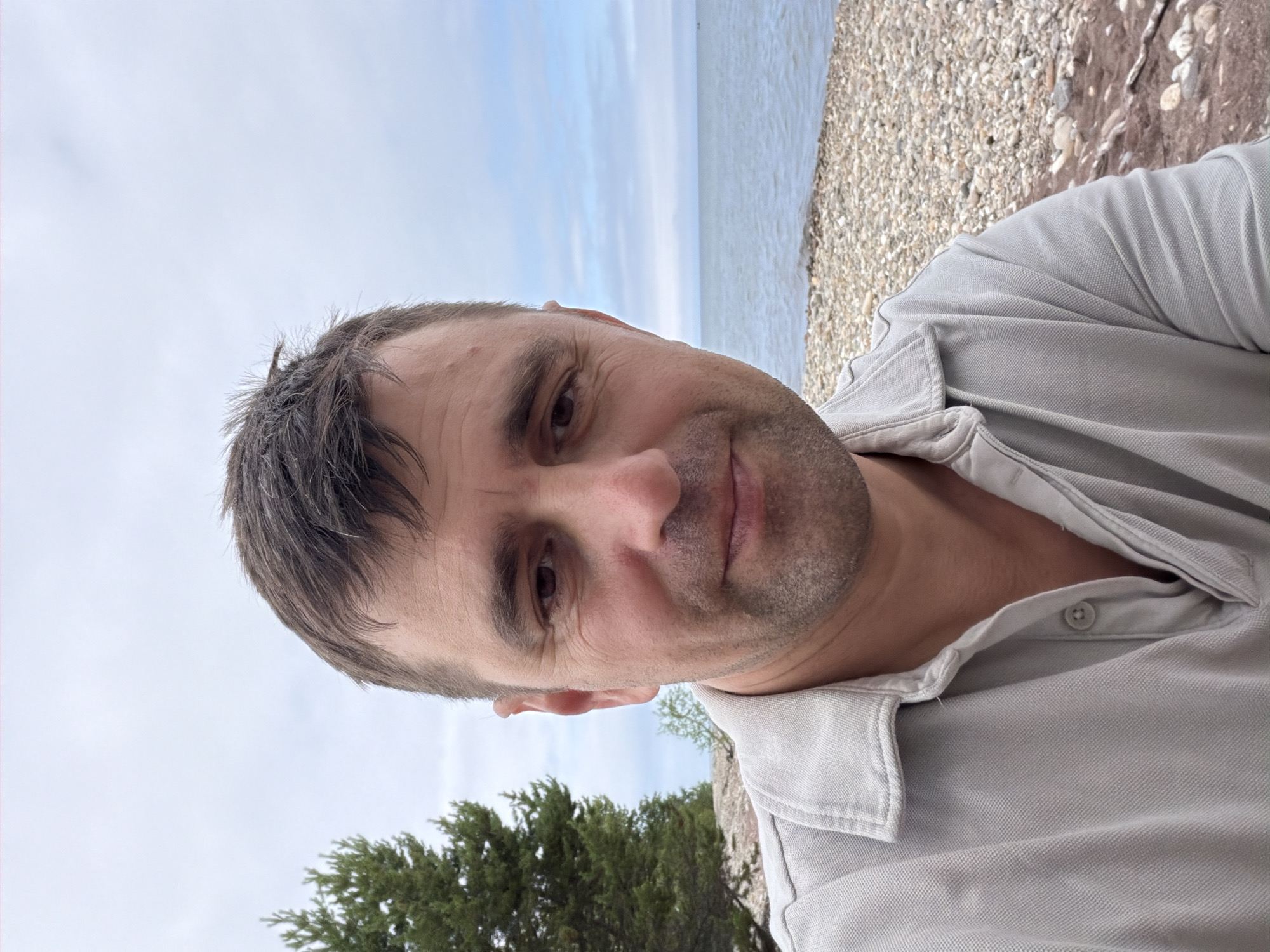 A man with short brown hair and stubble is taking a selfie outdoors on a cloudy day. He is wearing a light grey collared shirt. Behind him, a large green coniferous tree is on the left. Further back, there is a body of water with a shoreline covered in small stones and pebbles. The overall setting appears to be a natural environment, possibly a lakeside or coastal area near Utulik, Russia.