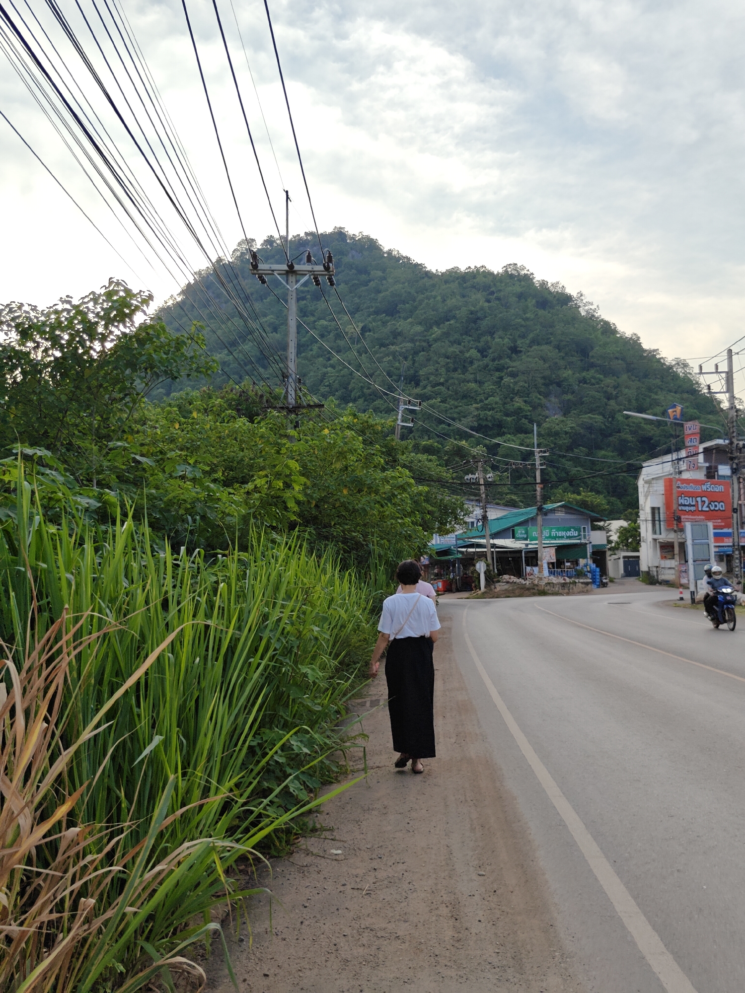 The image depicts a scene on a road in Mu Si, Thailand. A person, viewed from behind, walks along the edge of the road. They are wearing a white top and a long black skirt. Tall, green grass grows on the left side of the road, with some dried brown grass mixed in. To the right of the road, there are several buildings, including a store with a green awning and signage in Thai. A large, forested mountain rises in the background. Utility poles with numerous wires are visible throughout the scene. A motorcycle with two riders is seen on the road in the distance, moving away from the viewer. The sky is overcast.