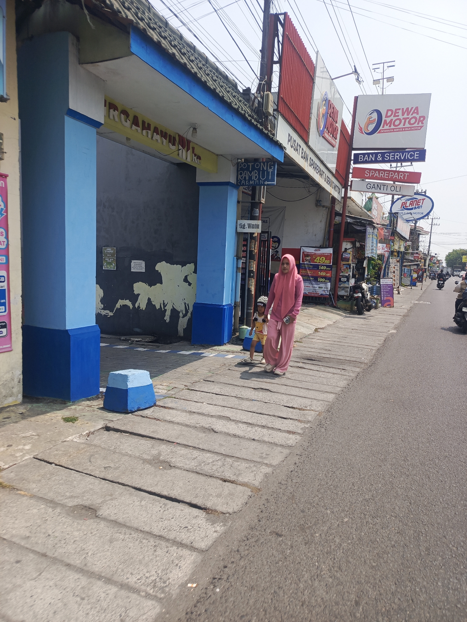 This is a street-level photograph taken outdoors on a sunny day in Sedati Gede, Indonesia. In the foreground, a concrete sidewalk runs parallel to an asphalt road. A woman wearing a pink hijab and matching pink pantsuit walks on the sidewalk, holding the hand of a small child wearing a white hat and a yellow shirt with a picture on it. The child is also wearing yellow shorts and white shoes.
To the left of the sidewalk, there is a building with blue and yellow painted columns framing an entrance. Above the entrance, a yellow sign with red lettering is partially visible, reading "RG AHAYU." Further to the right, a series of signs for businesses are visible. One prominent sign is white with red and blue lettering and reads "DEWA MOTOR" with the tagline "DEWANYA SERVIS & BAN MOTOR." Below this, there are smaller signs indicating "BAN & SERVICE," "SPAREPART," and "GANTI OLI." Another sign to the right reads "PLANET BAN MOTOR." A variety of motorcycles are parked along the side of the road and on the sidewalk. The road itself is populated with a few motorcycles and a car in the distance. Overhead, power lines are visible against a pale blue sky. The overall scene depicts a commercial street in an Indonesian town.