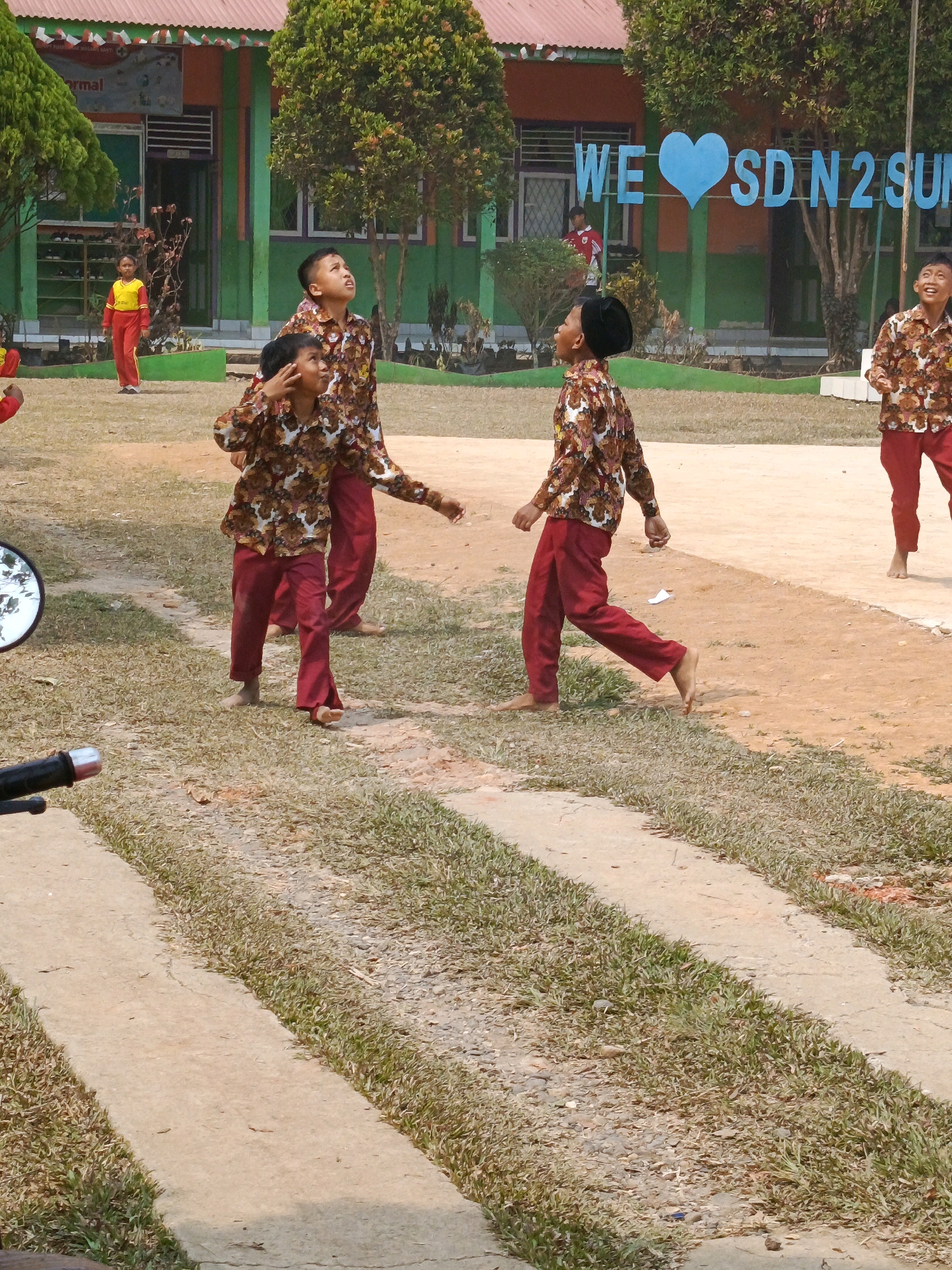 The image depicts an outdoor scene at what appears to be a school in Megang Sakti IV, Indonesia. In the foreground, a path of concrete runs through a grassy area. In the mid-ground, several young students in uniform are visible. The uniforms consist of patterned shirts and solid red pants. One student in the foreground is barefoot and appears to be walking or playing. Another student in the background is wearing a yellow and red uniform. Behind the students, a building with green and orange walls is visible, along with trees and a banner that reads "WE ♡ SD N 2 SUN." The overall environment suggests a bright, possibly warm day.