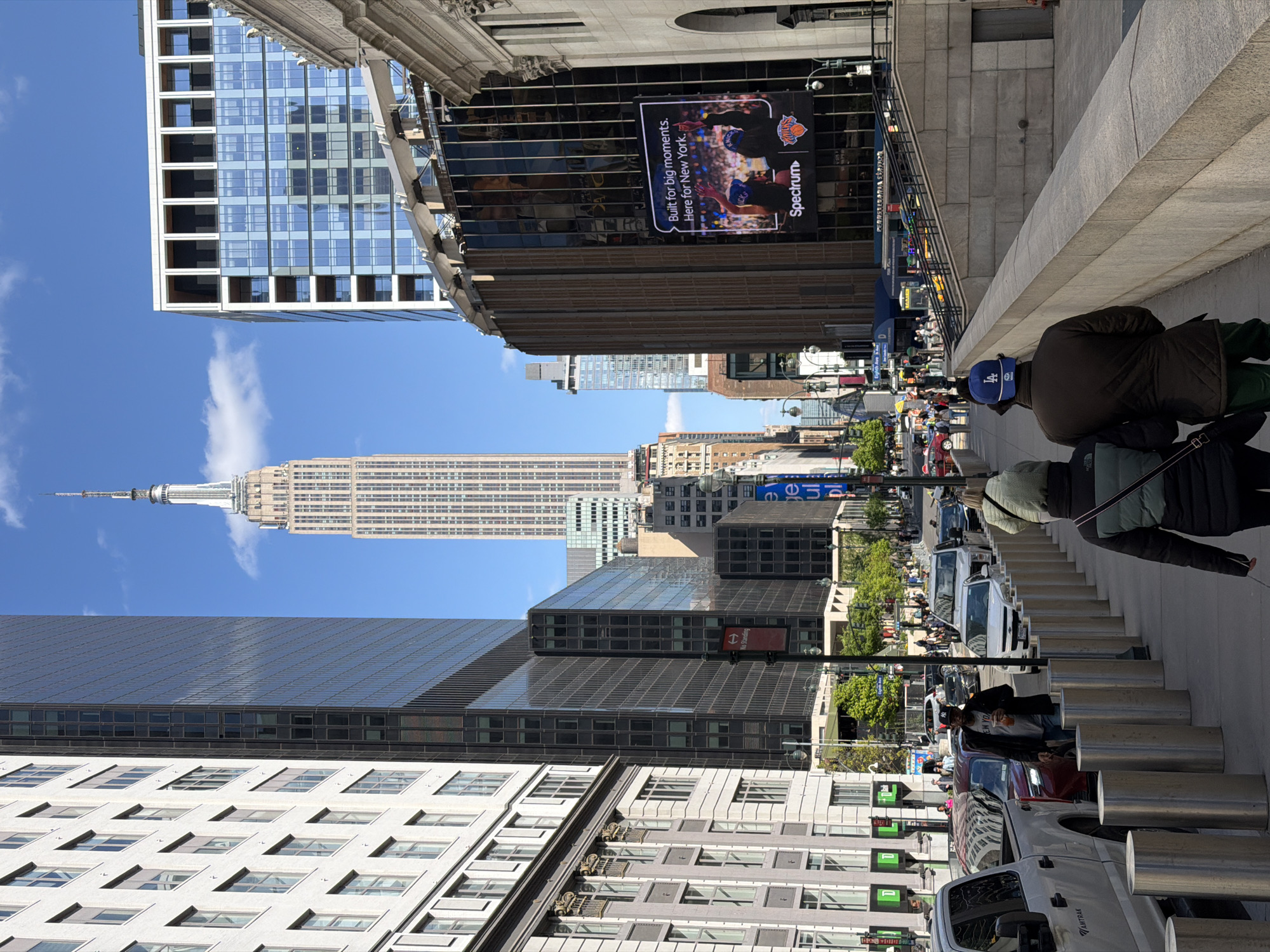 The image captures a street scene in New York City with the Empire State Building prominently visible in the background under a blue sky. The foreground includes various buildings, including a modern glass structure on the left and a building with an advertisement screen for Spectrum featuring the New York Knicks on the right. A line of silver bollards runs along the sidewalk, separating it from the street, where several vehicles are parked. People are walking on the sidewalk, one wearing a blue hat with an "LA" logo. Street signs and traffic lights are visible among the buildings, indicating a bustling urban environment.