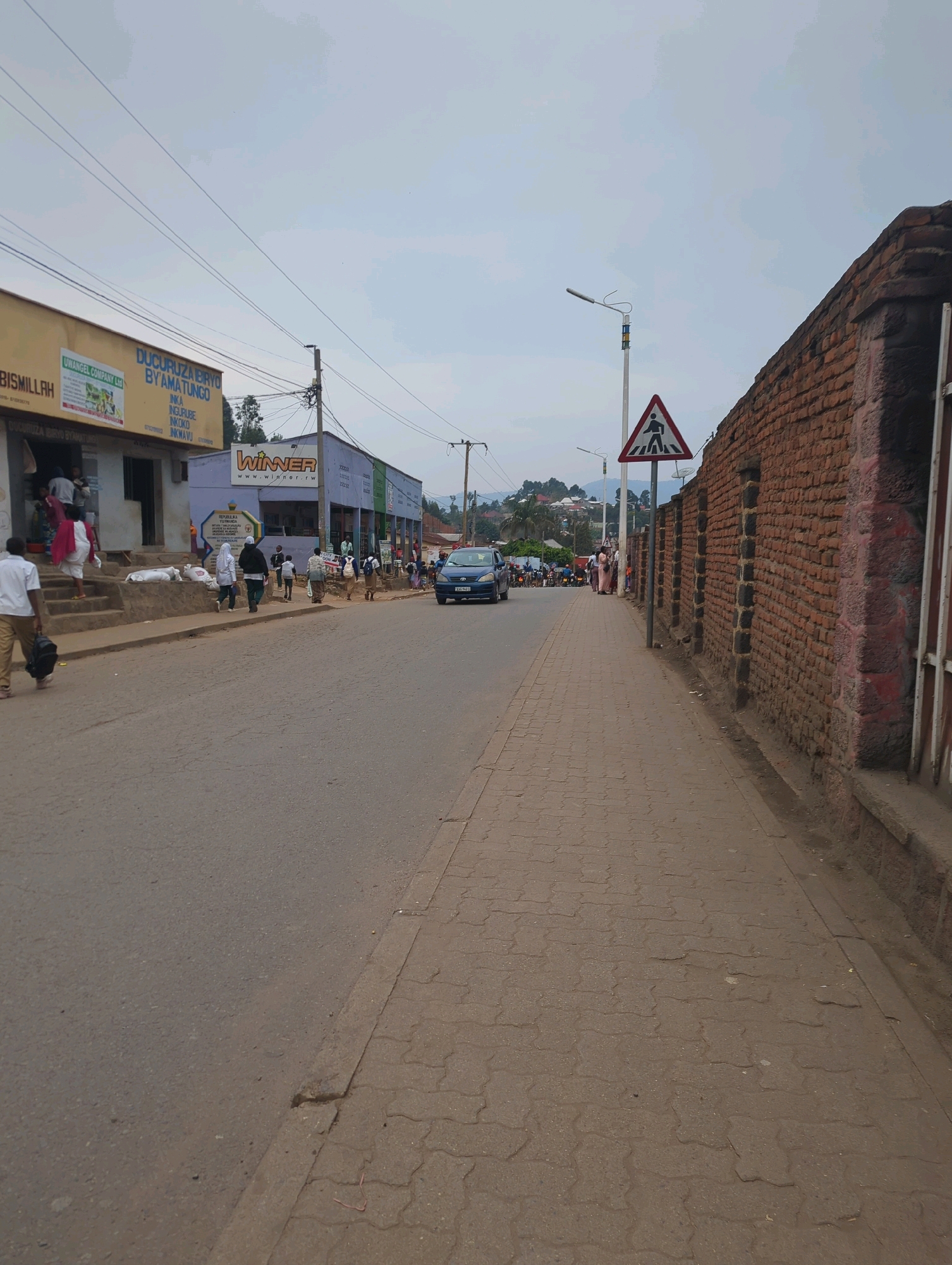 This is a street-level, eye-level, vertical, full shot of a street in Musanze District, Rwanda. The image captures a moderately busy street scene with several buildings lining the right side and a sidewalk. The buildings on the left appear to be shops, with awnings and signage. One sign reads "BISMILLAH" and another has "UWANGEL COMPANY LTD". Further down, a building with a large "WINNER" logo is visible. On the right side of the street, a long brick wall runs along the sidewalk, which is paved with interlocking bricks. A pedestrian crossing sign is visible on a pole. Several people are walking along the sidewalk and on the street. A blue car is driving on the road, and a few motorcycles are parked in the distance. The sky is overcast with a pale blue hue. The overall impression is that of a commercial area in a town or city.