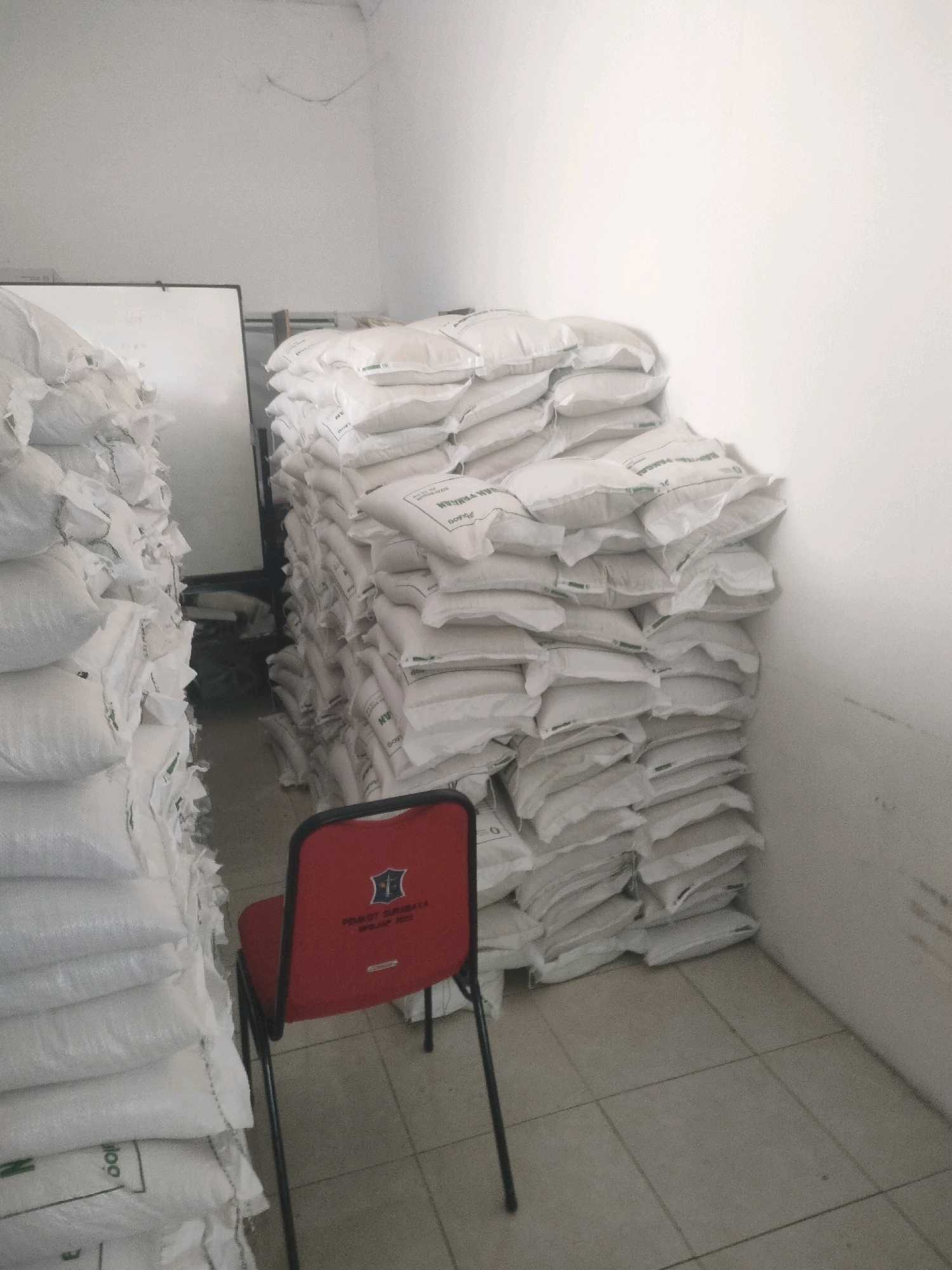 The image shows a storage room in Surabaya, Indonesia. Large stacks of white sacks are piled high against the walls. A red chair with black metal legs is placed in front of one of the stacks. The chair has a crest and text that reads "PEMKOT SURABAYA". The room has white walls and a tiled floor. A whiteboard is visible in the background. The lighting is dim and appears to be artificial.