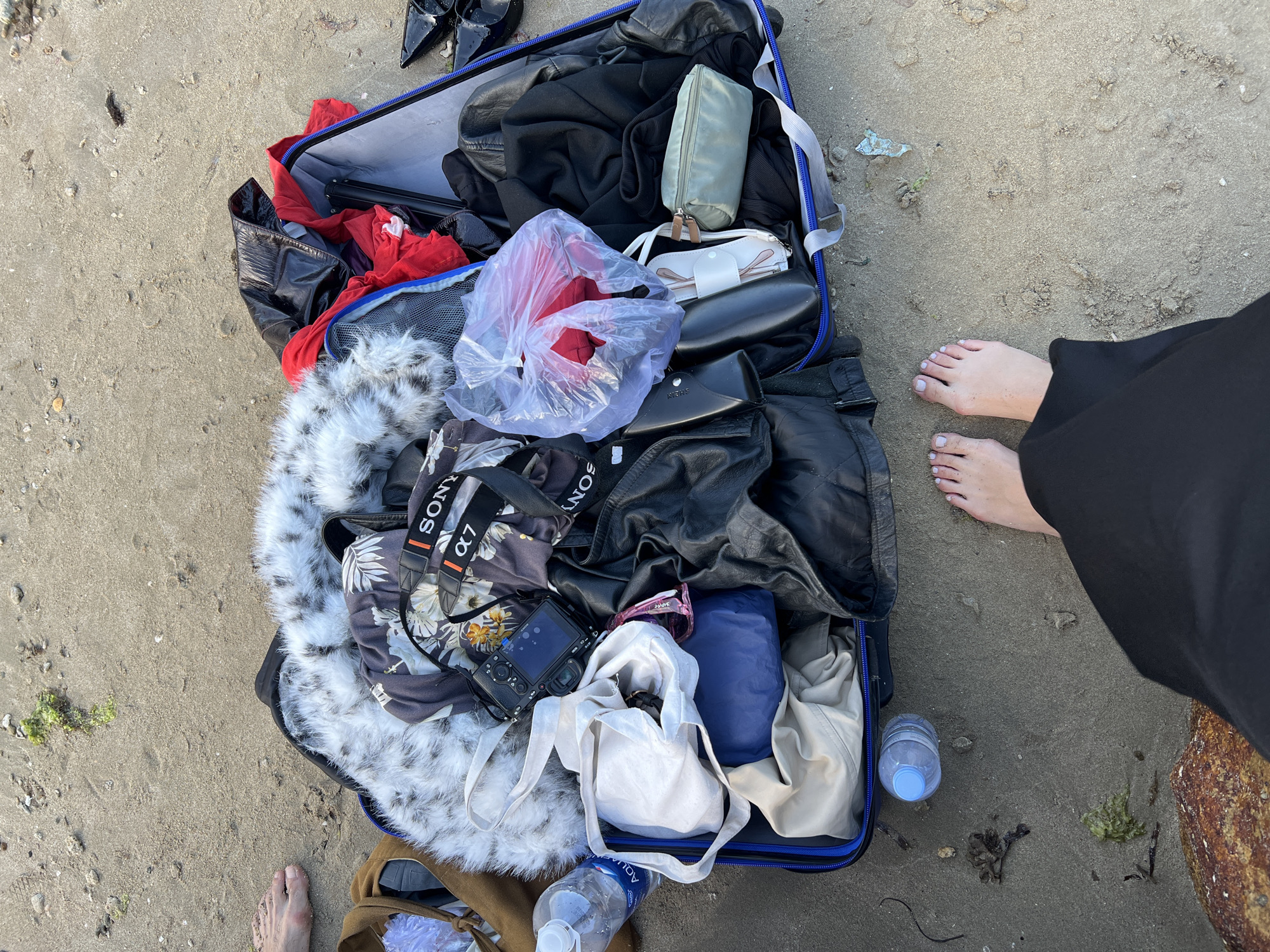 An overhead shot captures an open suitcase overflowing with various items on a sandy beach or coastal area in Bac Nha Trang Ward, Vietnam. The suitcase, with prominent blue trim, is packed with an assortment of clothing including black garments, red fabric (some in a clear plastic bag), a patterned floral shirt, a white furry item, and a beige jacket.
Personal items and accessories inside the suitcase include a light green rectangular pouch, a white eyewear case, a black sunglasses case, a black Sony α7 camera with its strap visible, a white canvas tote bag, a blue rolled item, and what appears to be a black tripod or stand.
Two individuals are present, evidenced by their bare feet on the sand. On the right, a person wearing long black clothing displays bare feet with light-colored nail polish. A second bare foot is partially visible at the bottom left, near brown-colored fabric.
The sandy ground around the suitcase contains scattered small debris, seaweed, and shell fragments. Two clear plastic water bottles with blue caps are visible, one partially empty and one full, with "AQUA" text discernible on the label of one. The scene is illuminated by natural daylight, suggesting it is daytime with pleasant weather. The arrangement suggests the individuals are either packing, unpacking, or sorting belongings on the beach.