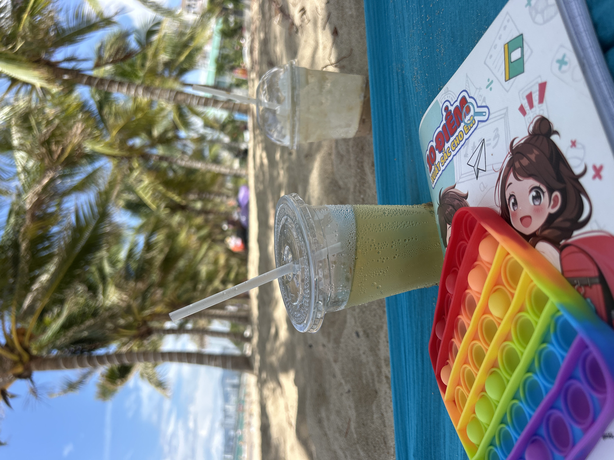 The image shows a beach scene in Nha Trang Ward, Vietnam. Two cups of drinks, one green and one white, are on a blue towel on the sandy beach. A notebook with an anime-style drawing and a rainbow-colored pop-it toy are also on the towel. In the background, palm trees are visible along the shoreline, and there is a glimpse of the ocean and the buildings of the city. The sky is blue with some clouds.