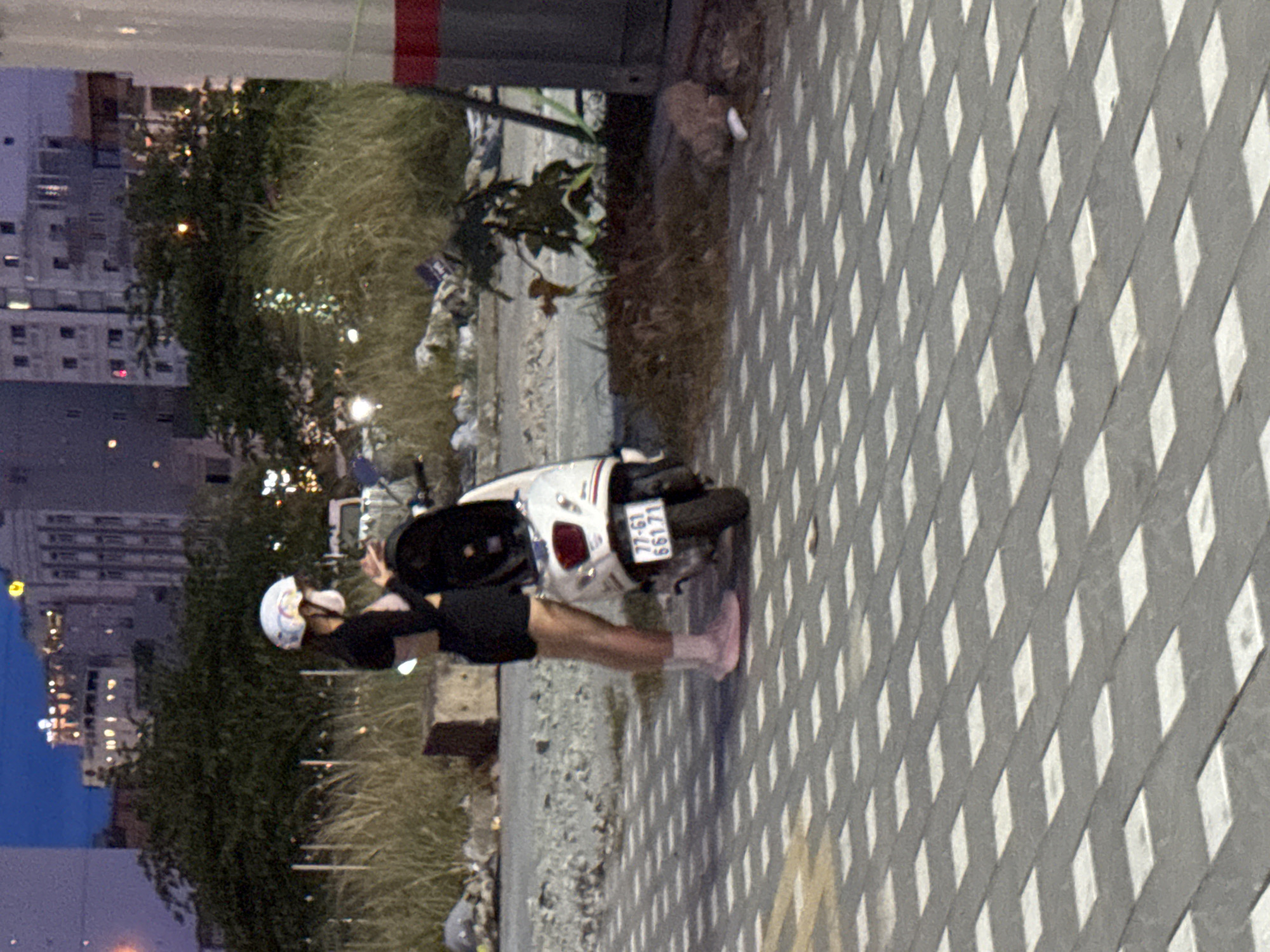 The image depicts a young woman standing next to a white scooter on a city street at dusk. She is wearing a black top, black shorts, pink socks, and pink shoes, and a white helmet with colorful decals. A white mask covers her nose and mouth. The scooter has a license plate with the characters "77-G1 661.71". The street is paved with light-colored square tiles, and shadows from overhead lights create a checkered pattern on the ground. In the background, there are tall buildings with illuminated windows, lush green trees, and sparse dry grass. The overall scene suggests an urban environment in Đà Nẵng, Vietnam, likely during an evening hour.