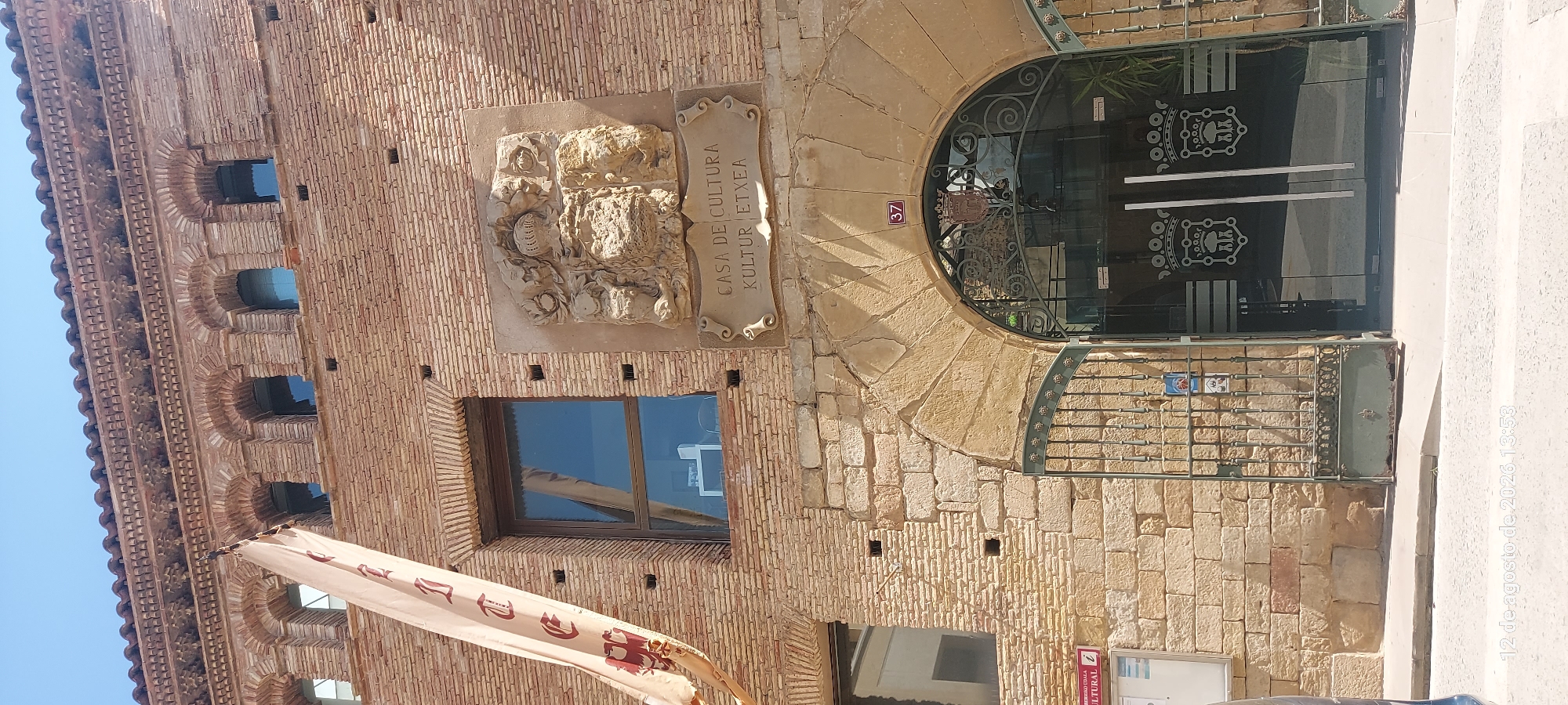 The image shows the exterior of the Casa de Cultura / Kultur Etxea in Olite / Erriberri, Spain. The building is constructed of reddish-brown brick with arched windows. A large stone relief of a coat of arms is mounted above an arched entrance, with a banner inscribed "CASA DE CULTURA / KULTUR ETXEA" below it. The entrance is a large glass door with metal gates in front of it. A sign with the number "37" is visible above the archway. To the left of the entrance, a notice board is mounted on the brick wall. A light-colored banner with dark lettering hangs diagonally from the upper left of the image, partially obscuring some of the upper windows. The sky is clear and blue. The scene is captured from a street-level perspective.