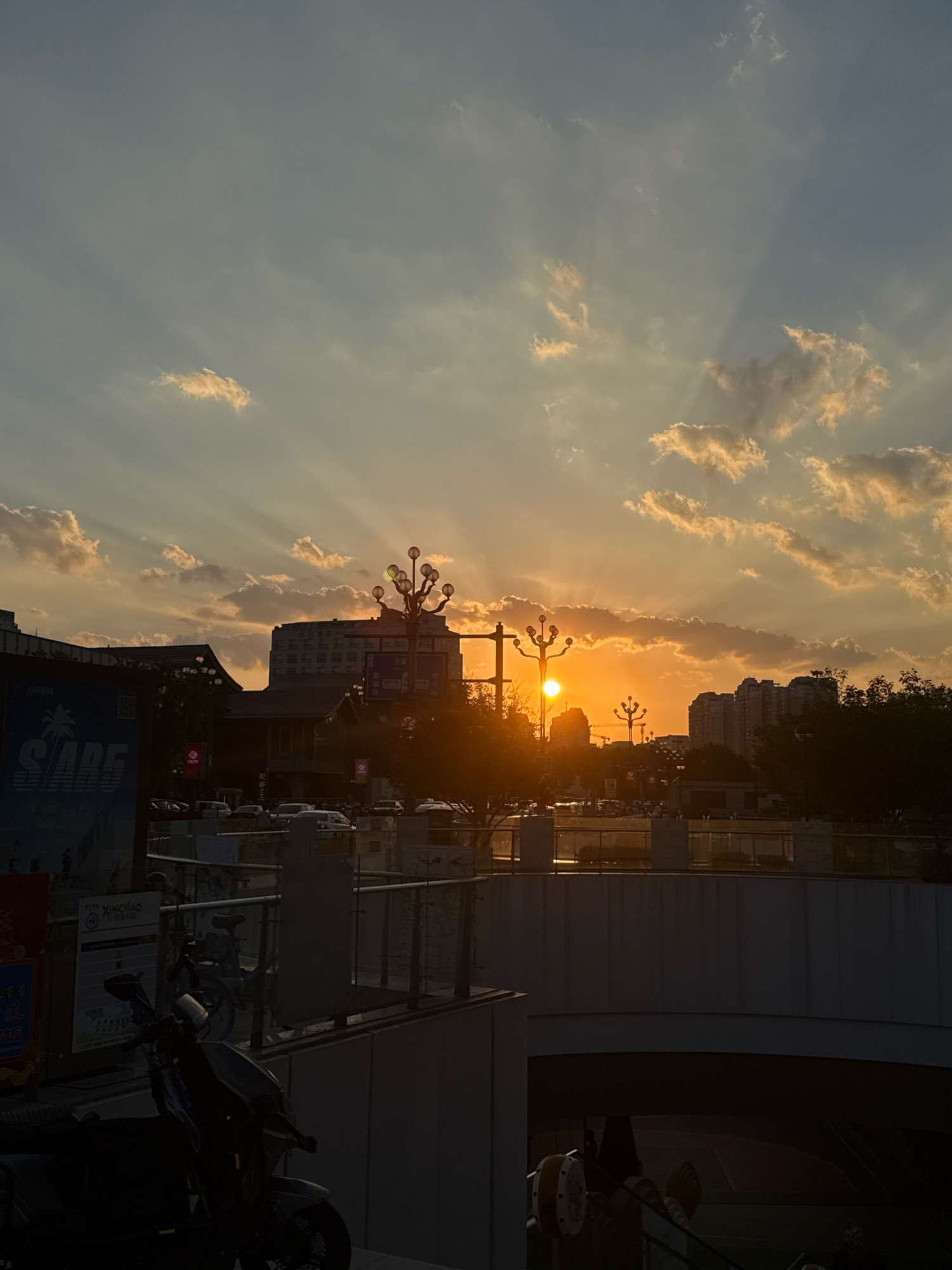 The image depicts a sunset scene in Yiwu, China. The sun is low in the sky, casting warm orange and yellow light through scattered clouds, creating visible sunbeams. In the foreground, a dark, silhouetted motorcycle is parked on a paved area. Above this, a raised walkway or platform is visible with railings and some parked bicycles. Beyond the platform, a street level view shows various buildings, including what appears to be commercial structures and residential high-rises. Several streetlights with multiple bulbs are also visible. Cars are parked and some are in motion on the street. The overall impression is a busy urban environment experiencing the end of the day.