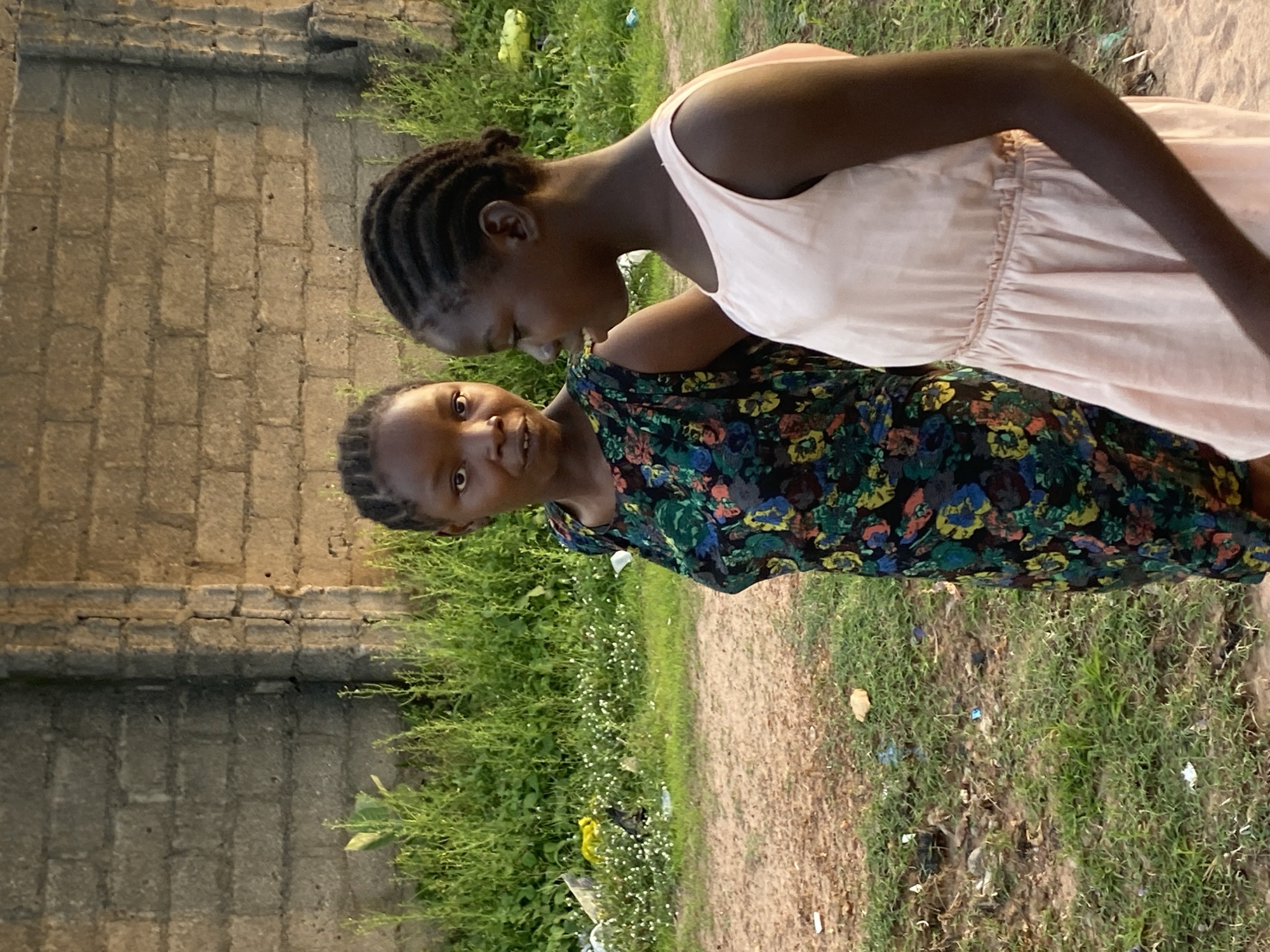 In Jauro Yinu, Nigeria, two young Black girls stand outside. The girl on the left wears a dark, floral-patterned dress and has her dark hair in braids. The girl on the right wears a light pink sleeveless dress and also has her dark hair in braids. They are standing in front of a textured, grey brick wall. In front of the wall is a patch of green grass with small white flowers and scattered debris. To the left of the girls is a dirt path. The lighting suggests it is either early morning or late afternoon.