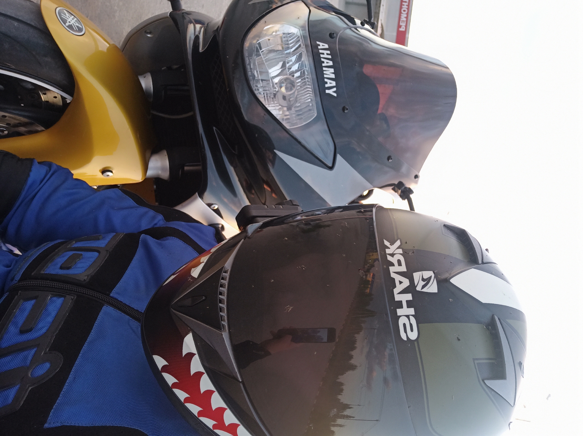The image shows a motorcycle helmet and the front fairings of two motorcycles. The helmet is black with white shark-like teeth painted on the front and a "SHARK" logo on the side. A gloved hand holding a phone taking the picture is reflected in the helmet's visor. To the left is the front fender of a yellow motorcycle. To the right is the front fairing of a black motorcycle with "AHAMAY" printed on it. The background is bright, suggesting an outdoor environment. The details needed to determine the city of Perm, Russia, are not apparent in the image.