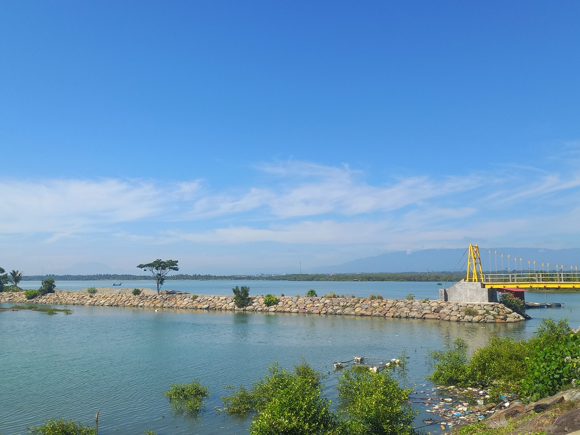 The image depicts a coastal scene in Lhokseumawe, Indonesia, on a clear day. A yellow suspension bridge extends over a body of water. A rocky barrier separates the foreground water from a larger body of water in the background. Green vegetation dots the landscape, including trees and shrubs. Some trash is visible in the water and along the shoreline. Mountains are faintly visible in the distant background under a blue sky with scattered clouds.