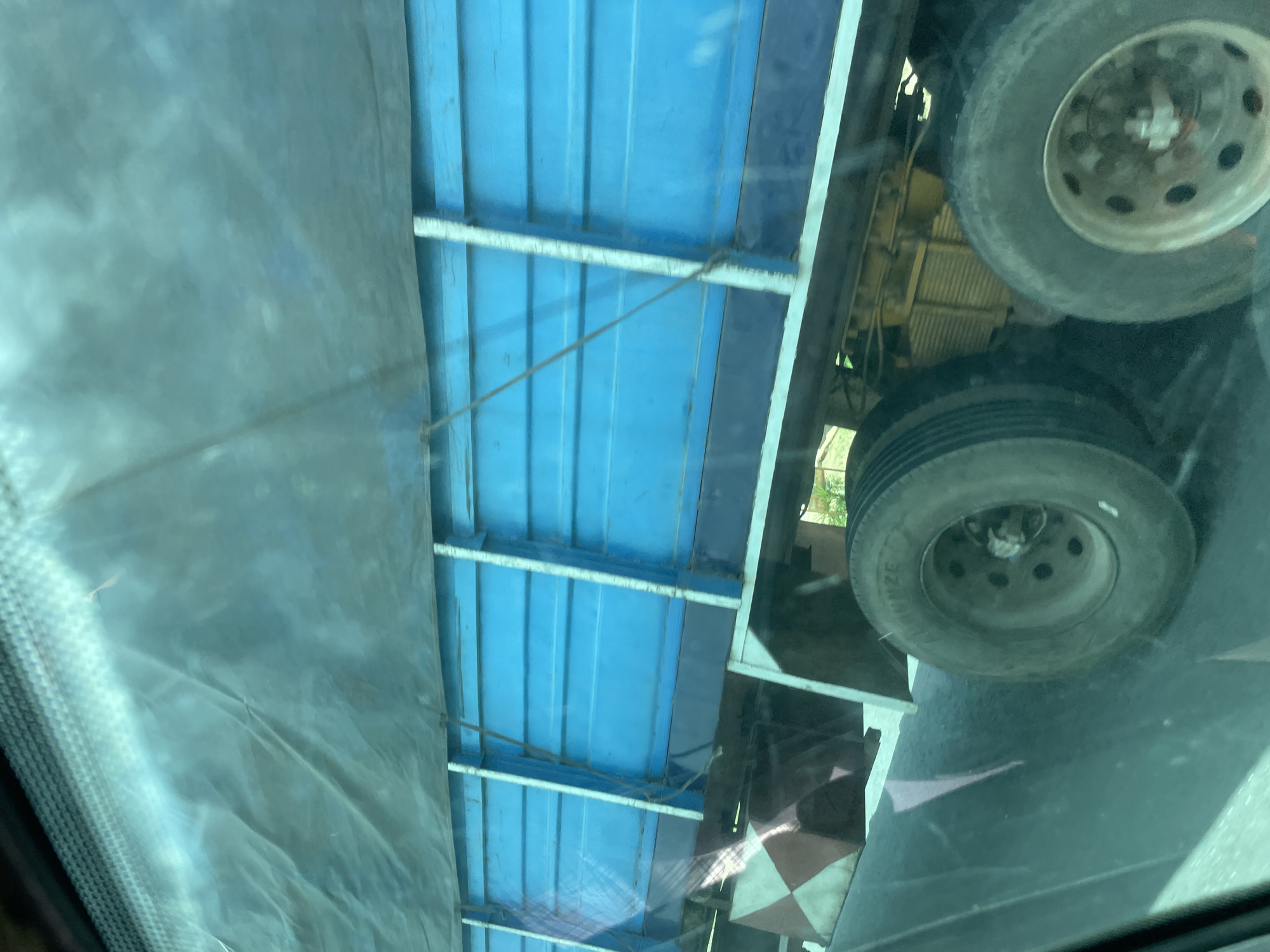 The image captures a side view of a large, blue truck with a tarp covering its cargo. The truck appears to be in motion, as suggested by the blurred background and the angle of the photograph, which seems to be taken from another vehicle. The truck has multiple wheels and a visible engine compartment. Reflective, diamond-shaped safety markings are present on the rear of the truck's bed. The scene is likely taking place on a road in Bauchi, Nigeria, given the context of the request. The environment appears to be somewhat bright and possibly sunny, with some vegetation visible in the background.