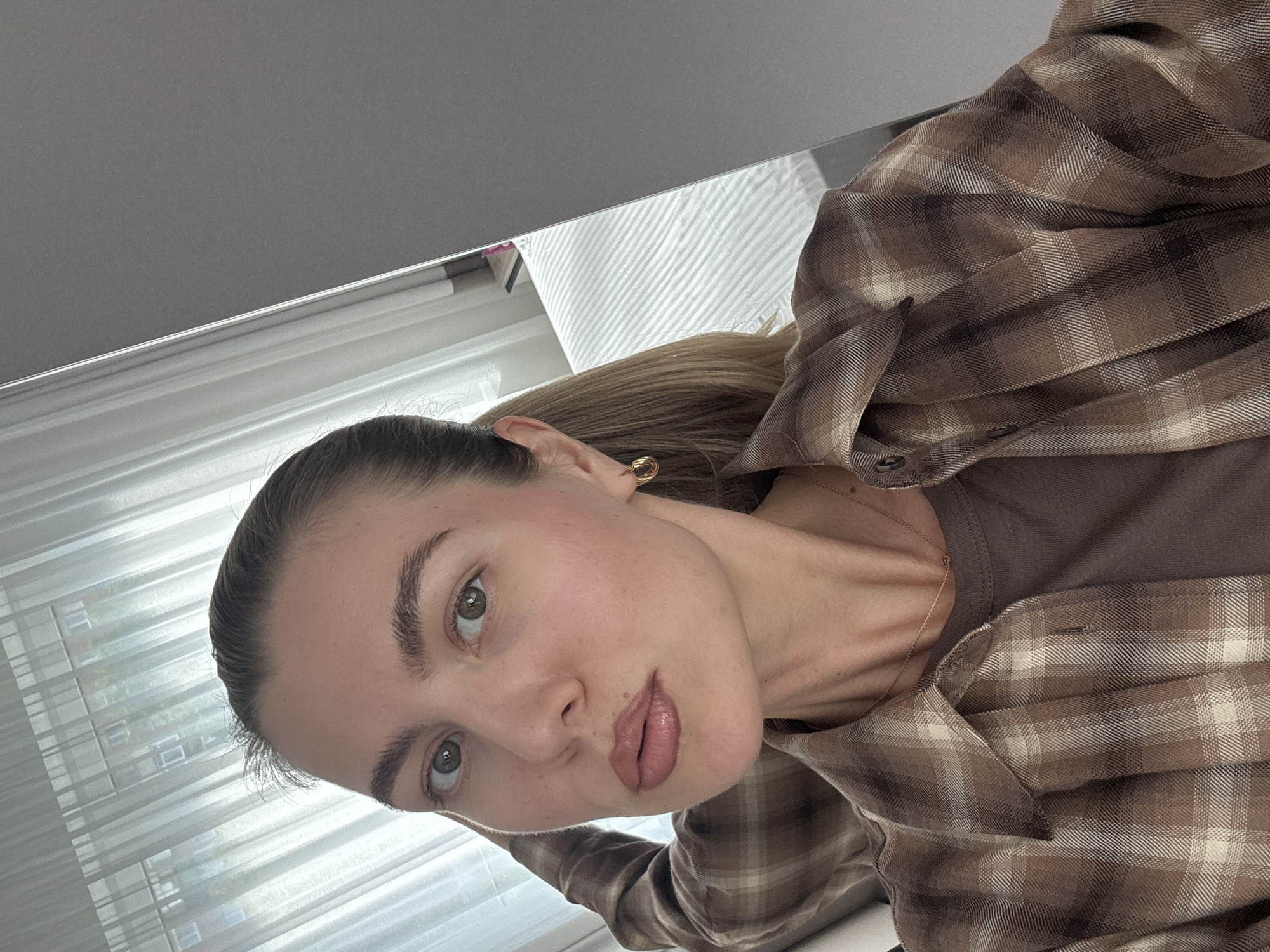 A close-up, low-angle selfie shows a young woman indoors, with the camera positioned above her head. She has light brown hair pulled back in a ponytail and is wearing a brown plaid button-up shirt over a brown t-shirt, with a delicate gold chain necklace. Her makeup is subtle, with a focus on rosy cheeks and a nude lipstick. She has green eyes and neatly shaped eyebrows. The background is blurred, featuring white vertical blinds that suggest a window or a partition. The lighting is soft and diffused, indicating it might be daytime. The overall mood is relaxed and casual.