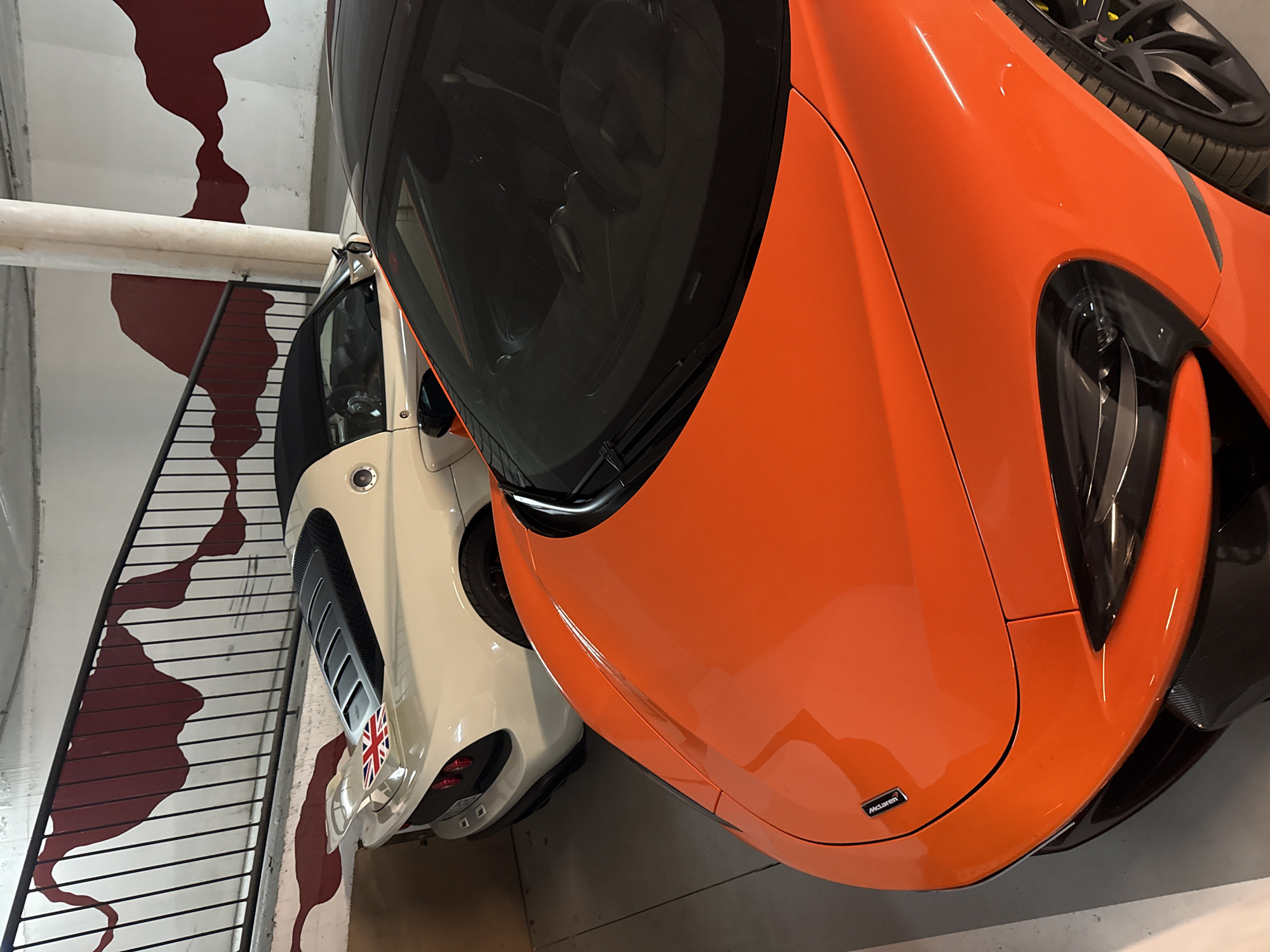 The image depicts two cars parked indoors, possibly in a garage or showroom. The foreground is dominated by an orange McLaren, with its front hood and windshield prominently visible. A "McLaren" logo is visible on the front. Next to the McLaren is a white car with a British flag emblem on the front, likely a Mini Cooper. The background includes a painted design on the wall, resembling a staircase with a red, abstract pattern alongside it. The overall setting seems to be a stylized or decorated parking area. The image does not provide explicit clues about Benasque, Spain.