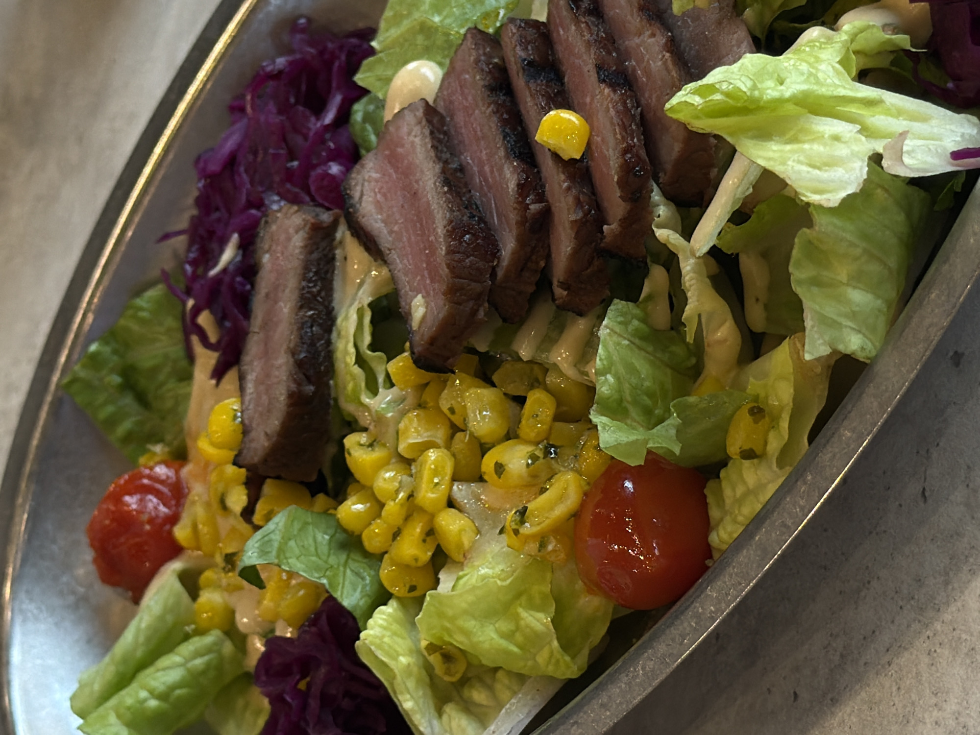 The image shows a close-up of a salad in a metallic dish, likely taken indoors. The salad ingredients include slices of seared steak, lettuce, corn, red cabbage, and cherry tomatoes. The location is unknown, and there is no information provided to suggest the location is Krasnoyarsk, Russia. No people are visible in the image.