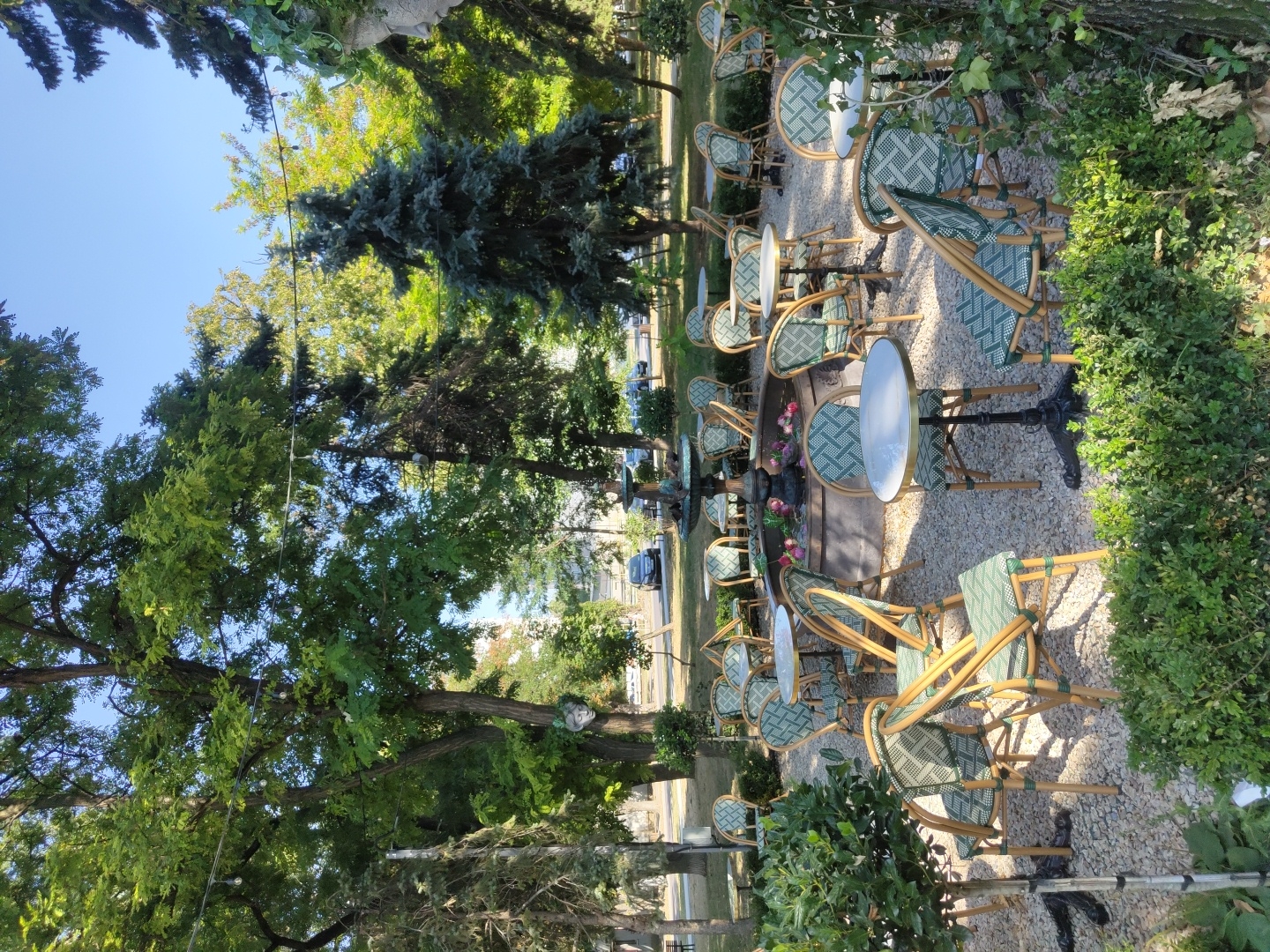 The image depicts an outdoor seating area of a cafe in Chișinău, Moldova, likely located in a park or garden setting, given the lush trees and greenery. The scene is bathed in bright sunlight, suggesting daytime. The area is furnished with numerous round tables and chairs, many with a distinctive green and white woven pattern on the seats and backs. A fountain is centrally located, surrounded by more seating. The ground is covered in light-colored gravel. In the background, trees line the perimeter, with some buildings and parked cars visible through the foliage, indicating proximity to a street or urban area. There are no people visible in the image.