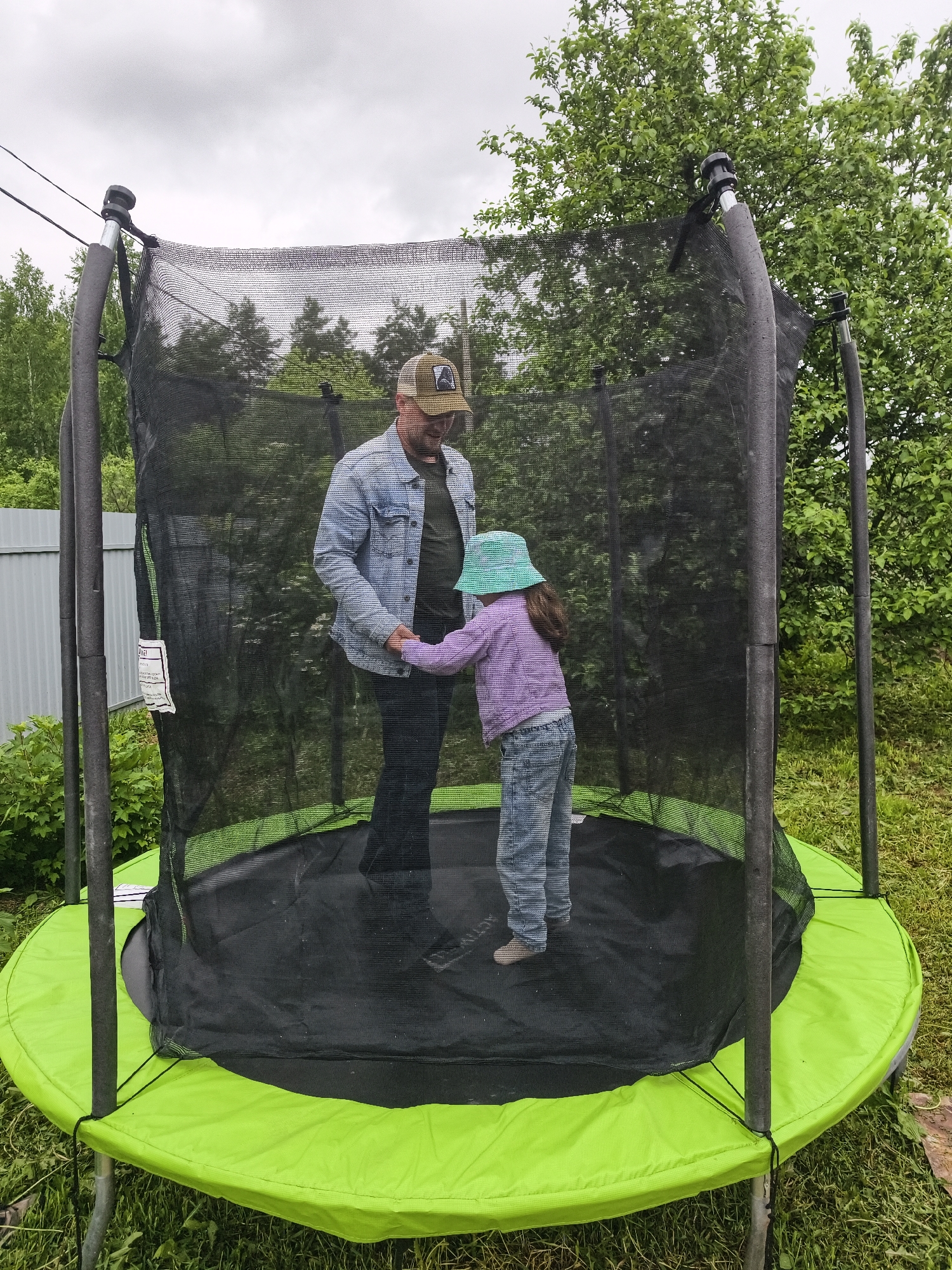 The image captures an outdoor scene, reportedly in the city of Prikamskii, Russia, featuring an adult male and a young child interacting within a trampoline. The environment is characterized by green foliage and an overcast sky.
The adult male stands on the left side of the trampoline's black jumping mat. He wears a light blue denim jacket over a dark t-shirt, dark trousers, and a khaki baseball cap featuring a dark emblem on the front. He has a light beard and is looking down at the child, smiling. His right hand holds the left hand of the child.
The young child is positioned to the right of the adult on the trampoline mat, with their back slightly turned towards the viewer. The child wears a light blue bucket hat, a purple long-sleeved top, light-wash blue jeans, and light-colored footwear. Their right hand holds the adult's left hand.
The trampoline is circular, with a black jumping surface and bright lime-green protective padding covering the springs and frame. A black safety net encloses the perimeter, supported by dark, foam-padded poles. A white instructional or warning tag is visible on the safety net on the left side of the frame.
The surrounding landscape consists of green grass on the ground. In the background to the left, a grey corrugated metal fence is visible, with distant green trees beyond. To the right, a dense canopy of lush green deciduous trees forms the primary background. The sky above is uniformly grey and overcast.