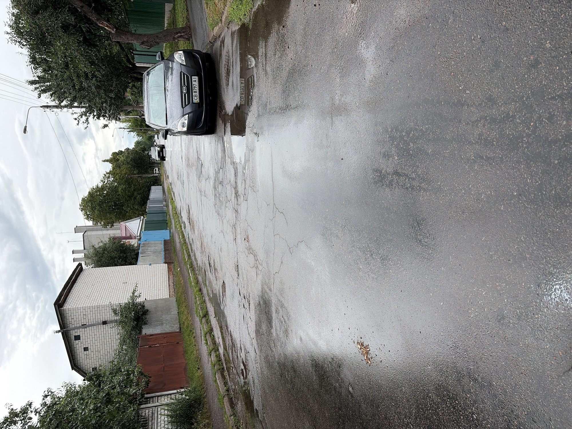 The image depicts a street scene in Chernihiv, Ukraine, likely after rainfall, as evidenced by the wet asphalt and puddles. A dark gray Kia car, with license plate CB 8797 AI, is parked on the right side of the road, facing the viewer. The road itself is in a state of disrepair, with visible cracks and potholes, further indicating the presence of water.
To the left of the road, a sidewalk is visible, bordered by a curb and grass. Beyond the sidewalk, there are fences, including a rusty metal fence and fences painted in blue and green. A white brick building with a dark roof and several chimneys is situated behind the fences.
On the right side of the road, beyond the parked car, there is a green fence and lush green trees with dark foliage. The sky is overcast with a cloudy, gray appearance. The overall environment suggests a residential or semi-industrial area within the city. No people are visible in the image.