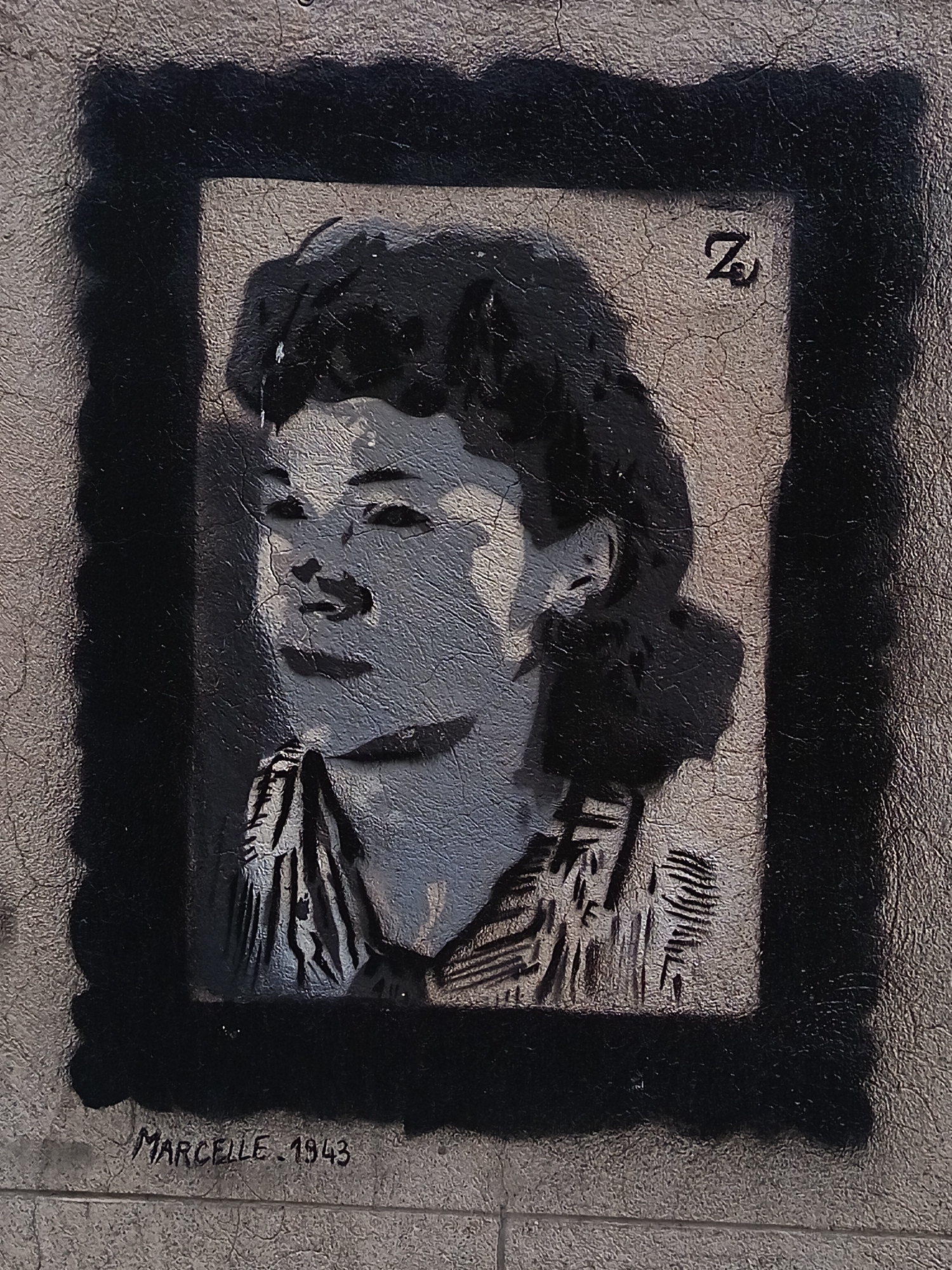 This is a stencil graffiti artwork on a textured concrete wall, depicting a portrait of a young woman. The portrait is rendered in black and gray tones and is framed by a thick, uneven black border. In the upper right corner of the frame, the letter "Z" is stenciled. Below the portrait, the name "MARCELLE" and the year "1943" are written in a handwritten script. This artwork is located on the wall of a building in the city of Périgueux, France, specifically on Rue de la Clarté, near the former Jewish ghetto. The woman in the portrait is likely a reference to Marcelle, a victim of the Holocaust. The overall condition of the artwork is good, with some visible cracking in the concrete wall beneath the paint.