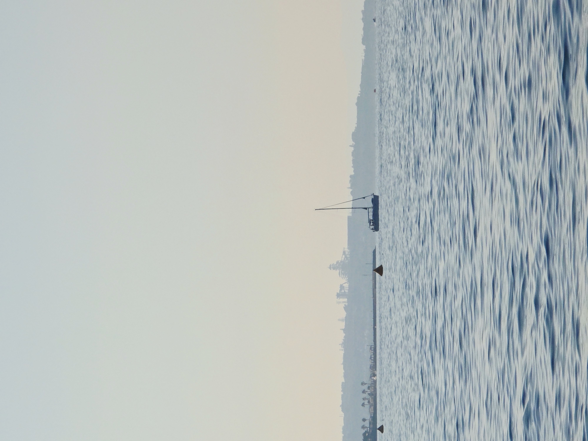 The image depicts a serene coastal scene with calm, blue waters. In the middle ground, a sailboat with a single mast navigates the sea. The distant shoreline features a hazy silhouette of buildings and what appears to be an amusement park or resort with tall structures. Palm trees are visible along the coast, suggesting a warm climate. Two orange buoys are sparsely scattered in the water, marking an area. The sky is a pale, hazy blue, indicating soft lighting conditions, possibly early morning or late afternoon. The overall impression is one of tranquility and a coastal landscape.